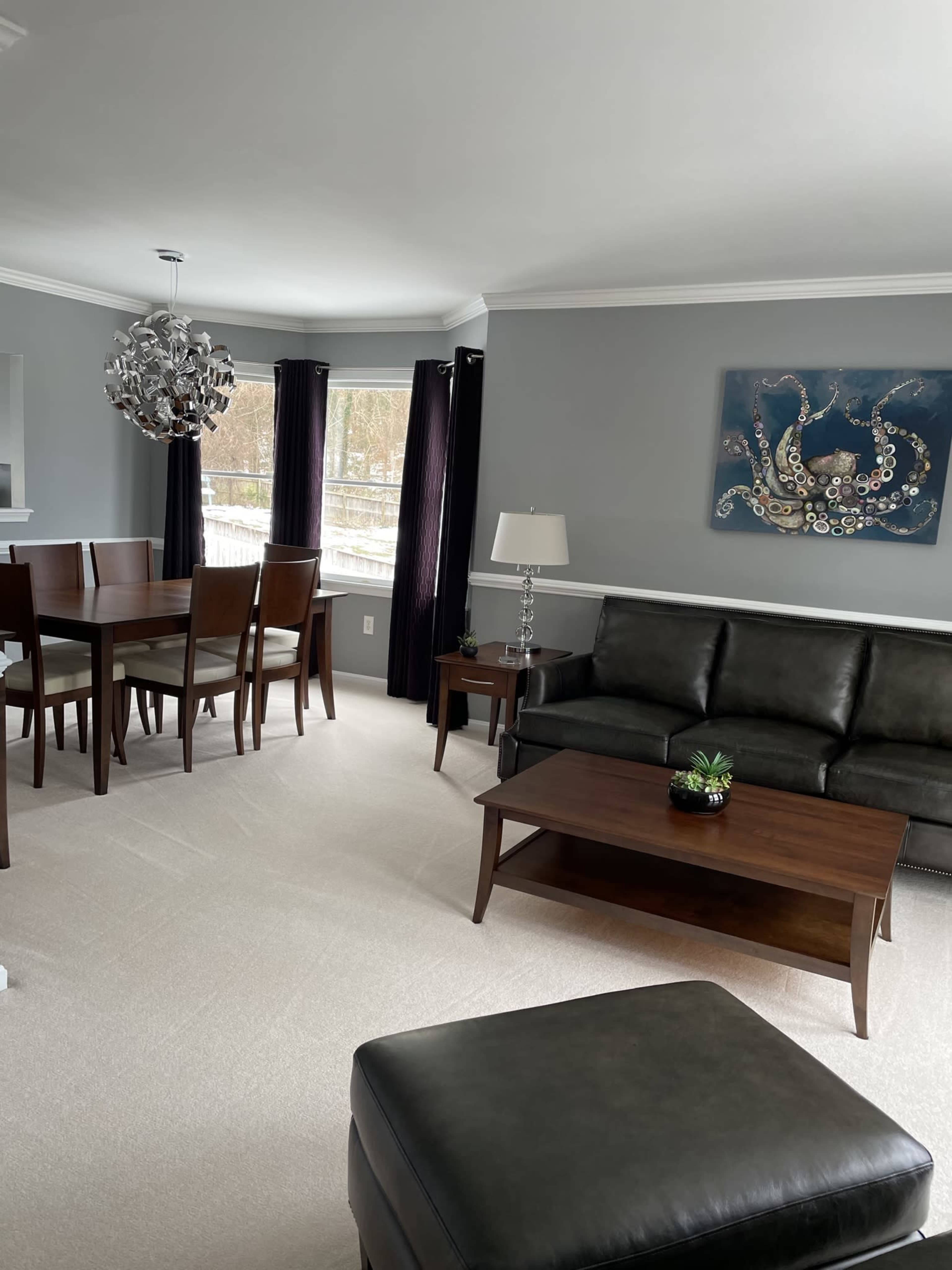 A modern living room with a dark leather sofa, a wooden coffee table, a dining area with a table and chairs, and large windows covered by black curtains.