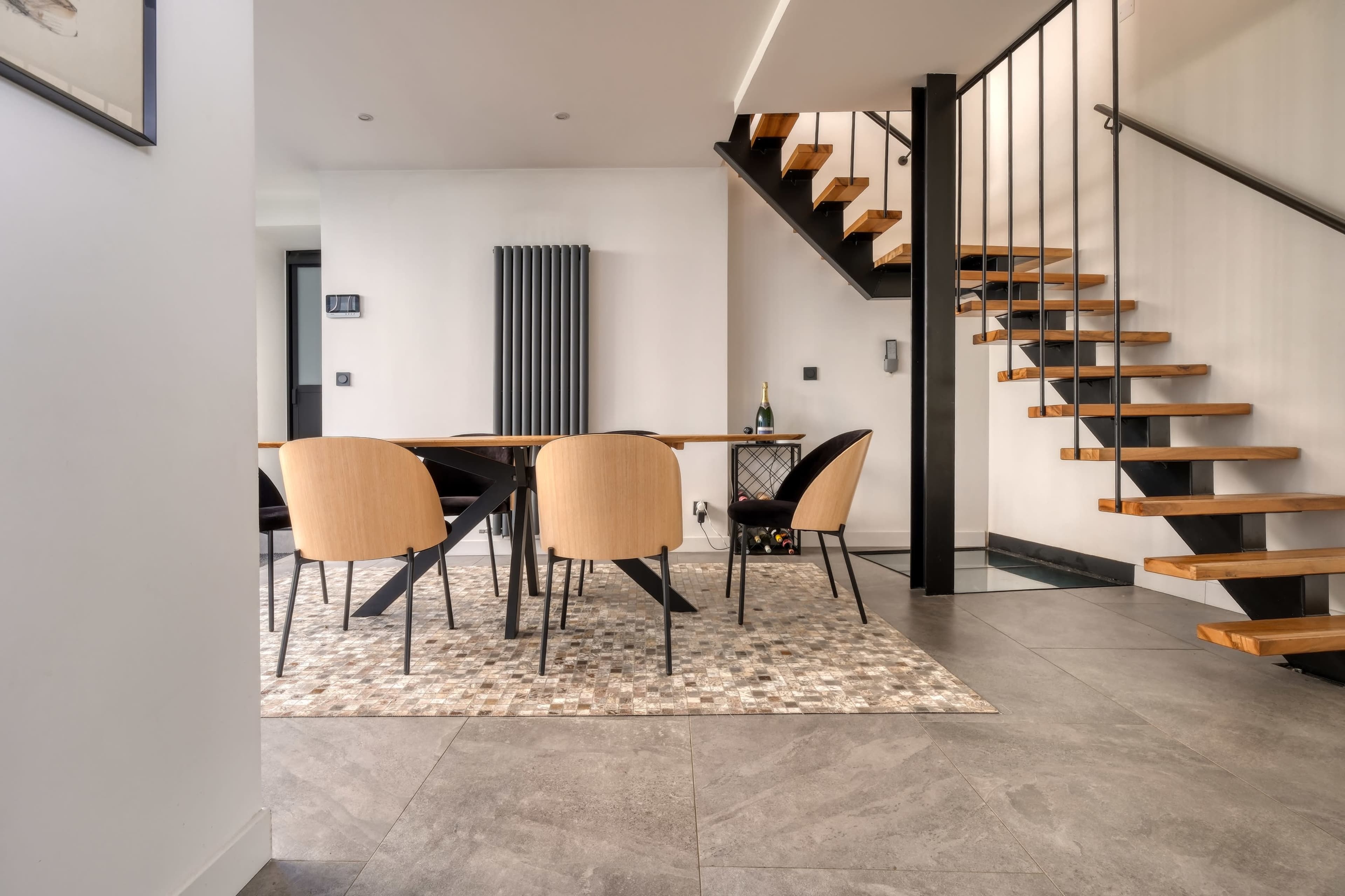 The image shows a modern interior space featuring a dining area with a table and chairs, a staircase, and large tiles on the floor.