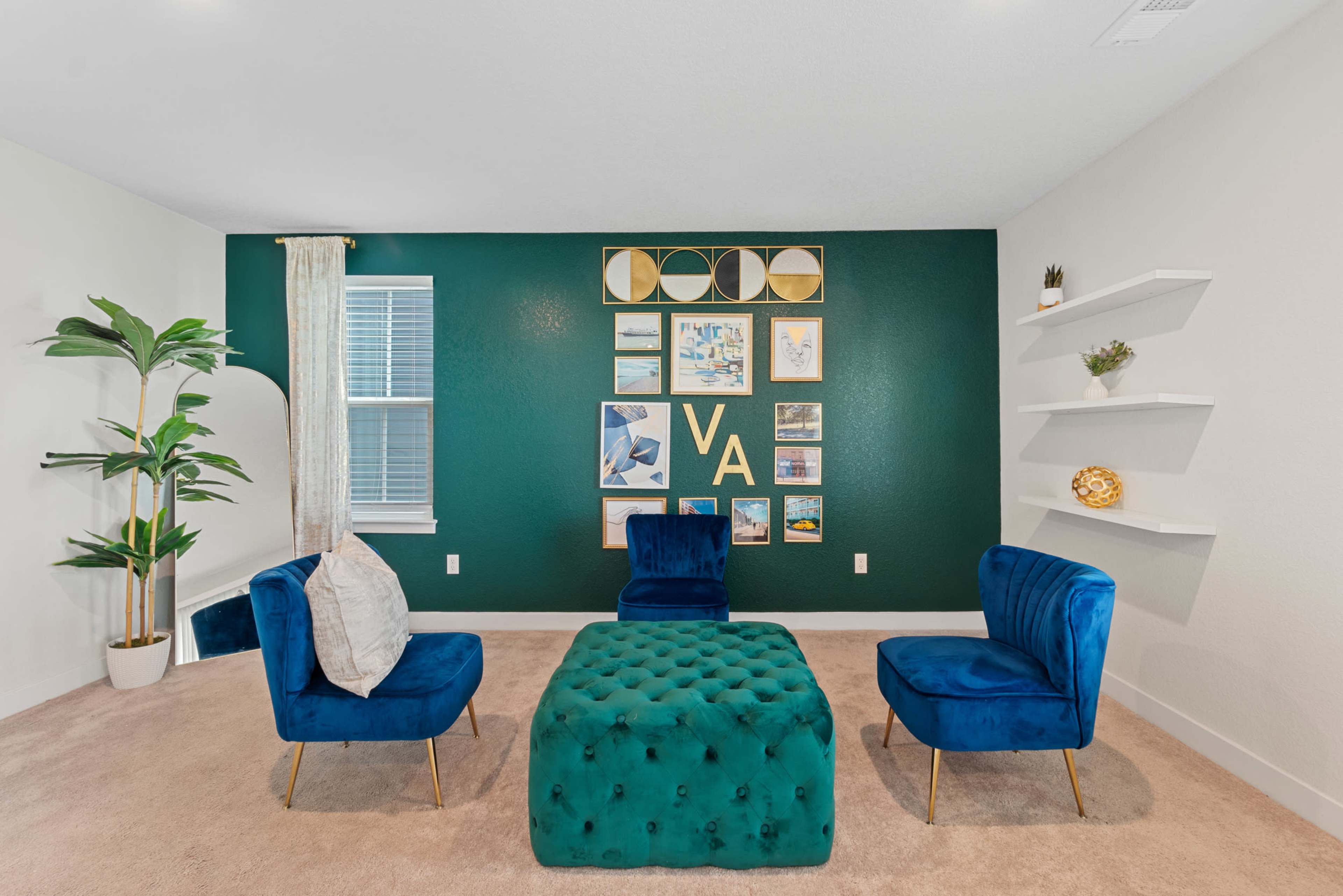 A cozy room features two blue chairs and a tufted green ottoman, with a green accent wall adorned by framed artwork.
