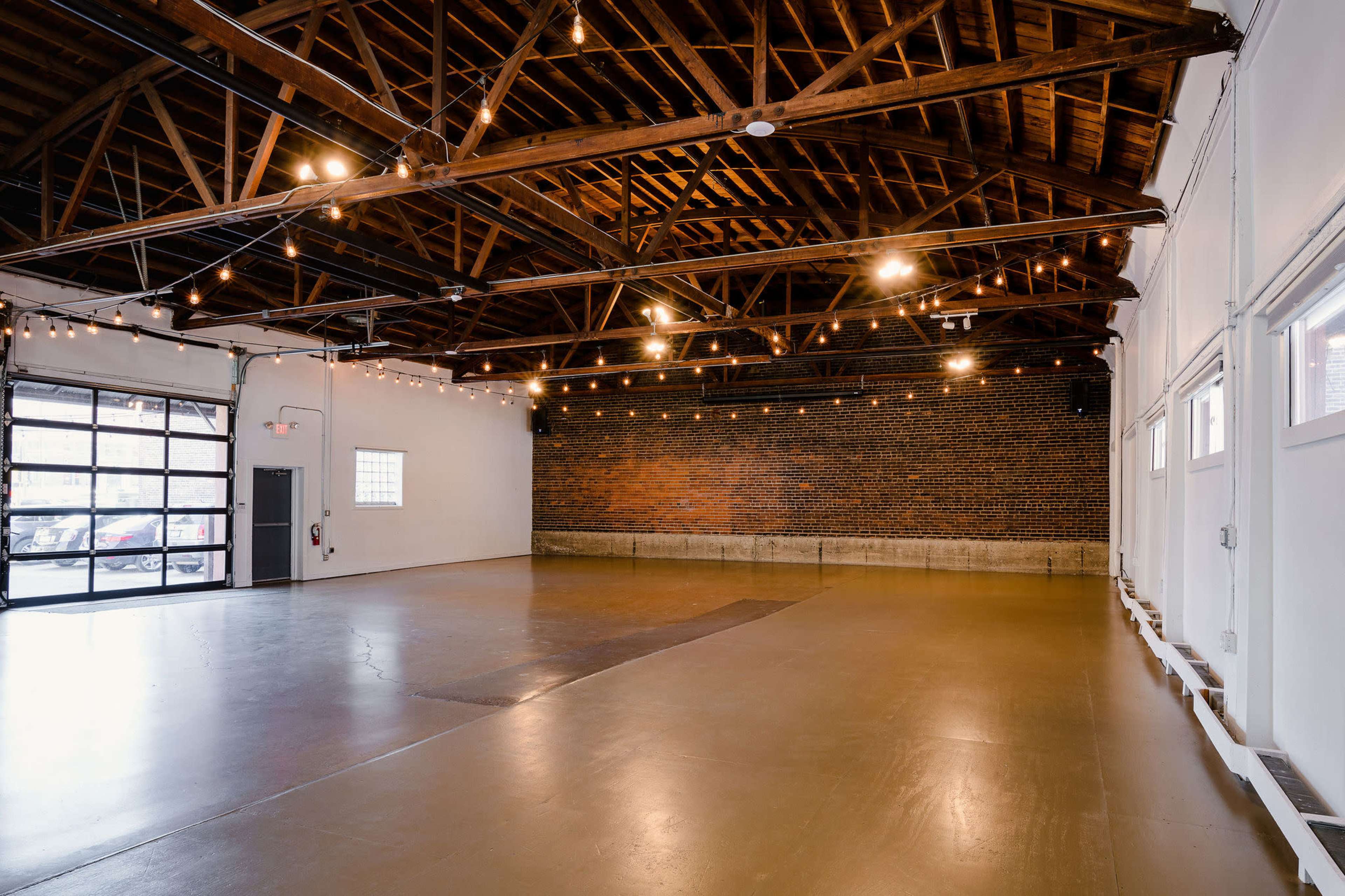 A spacious, industrial-style room with exposed wooden beams, string lights, and a brick wall, featuring a polished concrete floor and large garage doors.