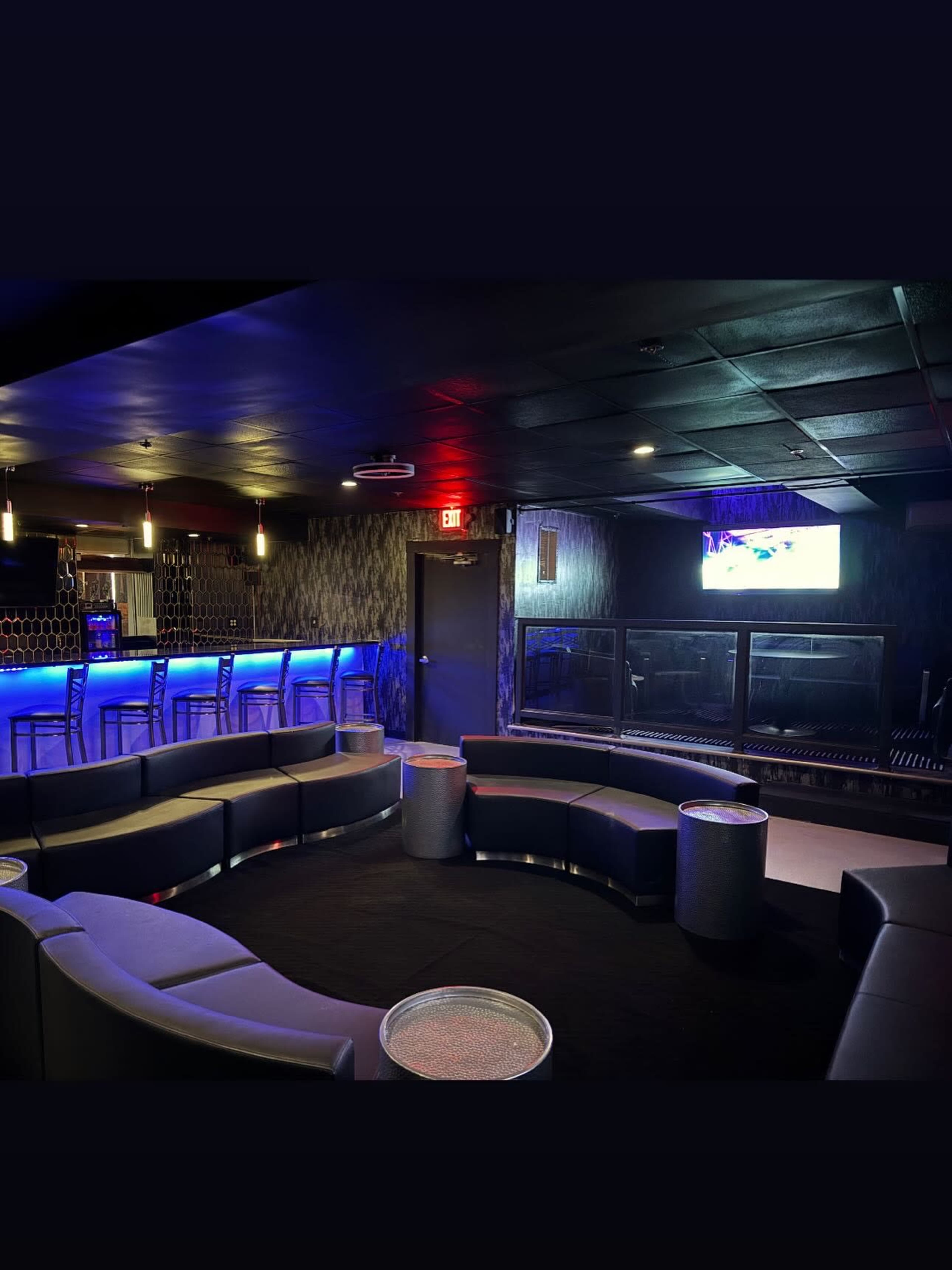 The image shows a modern lounge with curved seating arrangements, ambient lighting, a bar area, and a television mounted on the wall.