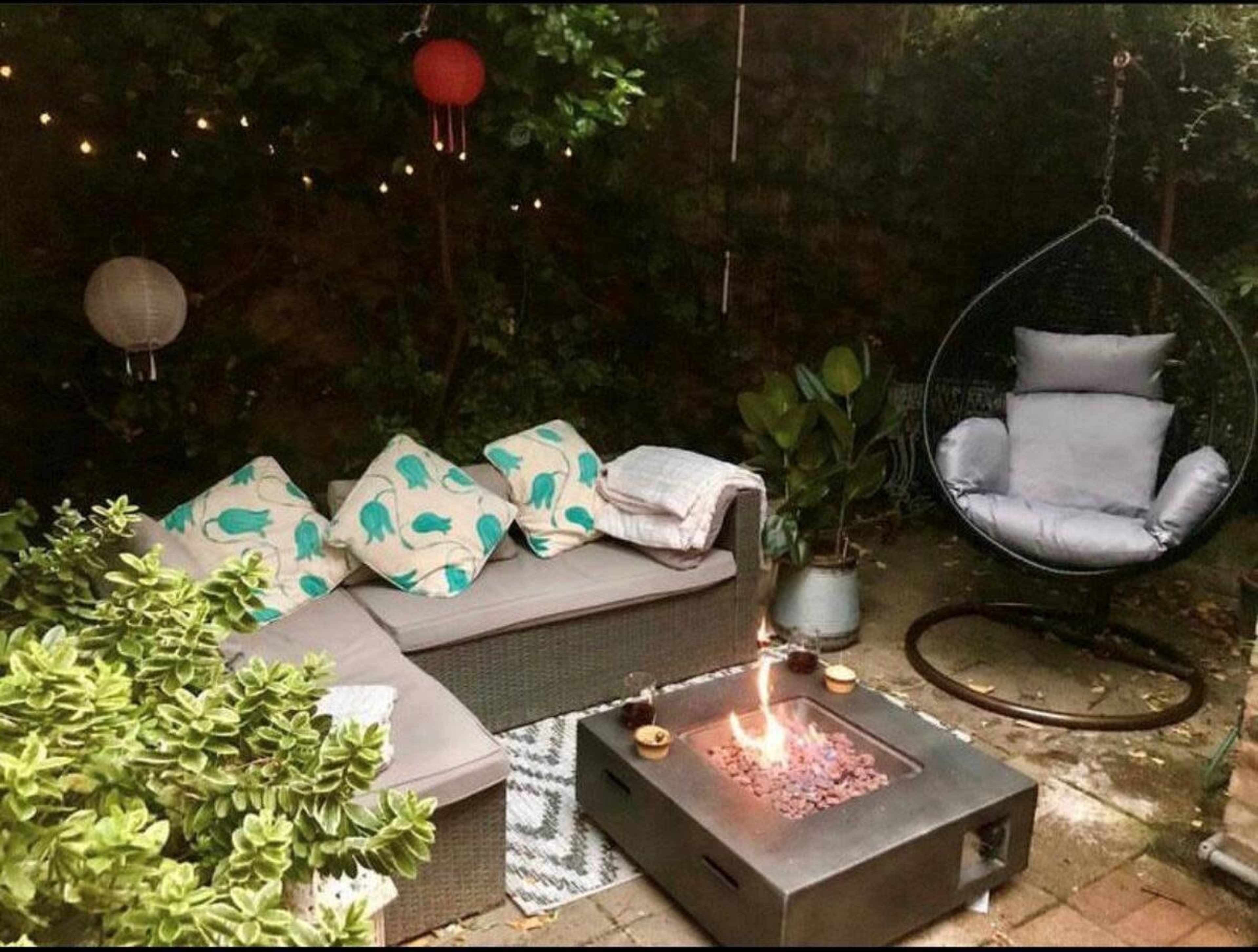 A cozy outdoor seating area with a sectional sofa, hanging chair, and a fire pit surrounded by green plants and decorative lanterns.
