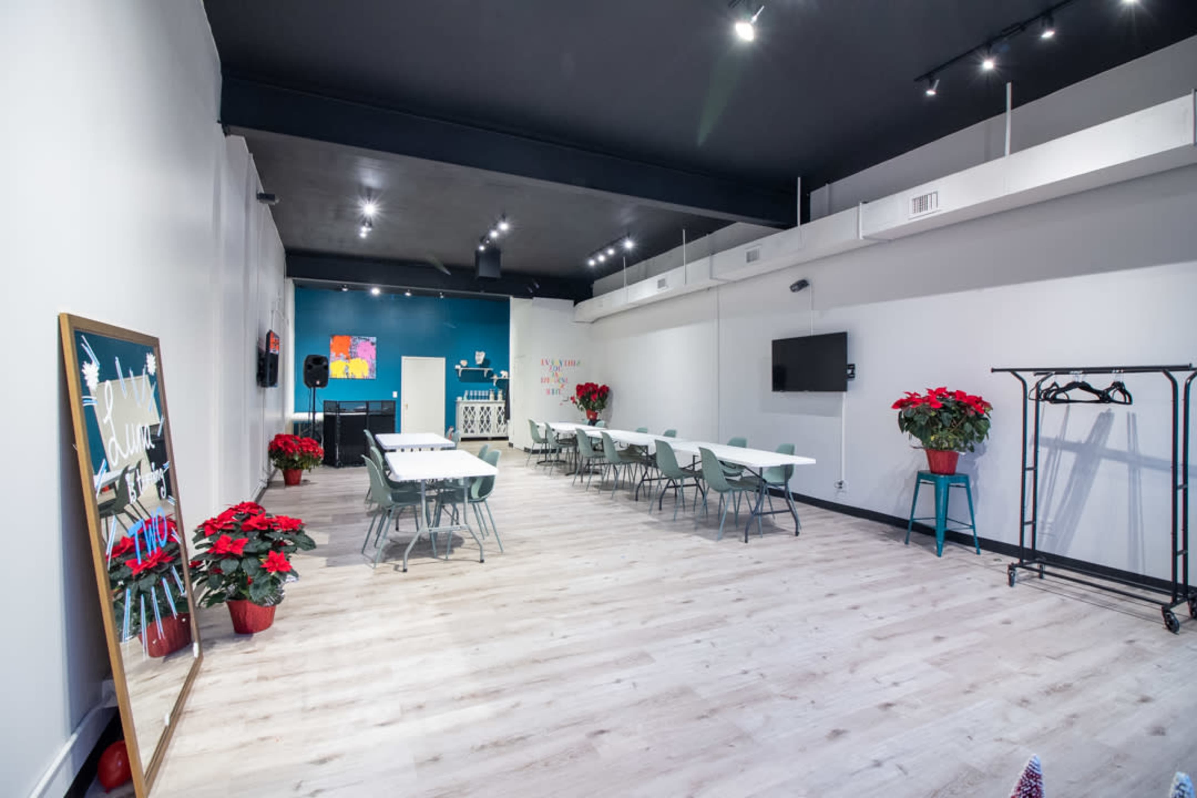 A spacious room with light wooden flooring, tables arranged for seating, potted poinsettias, and a wall with a mounted screen.