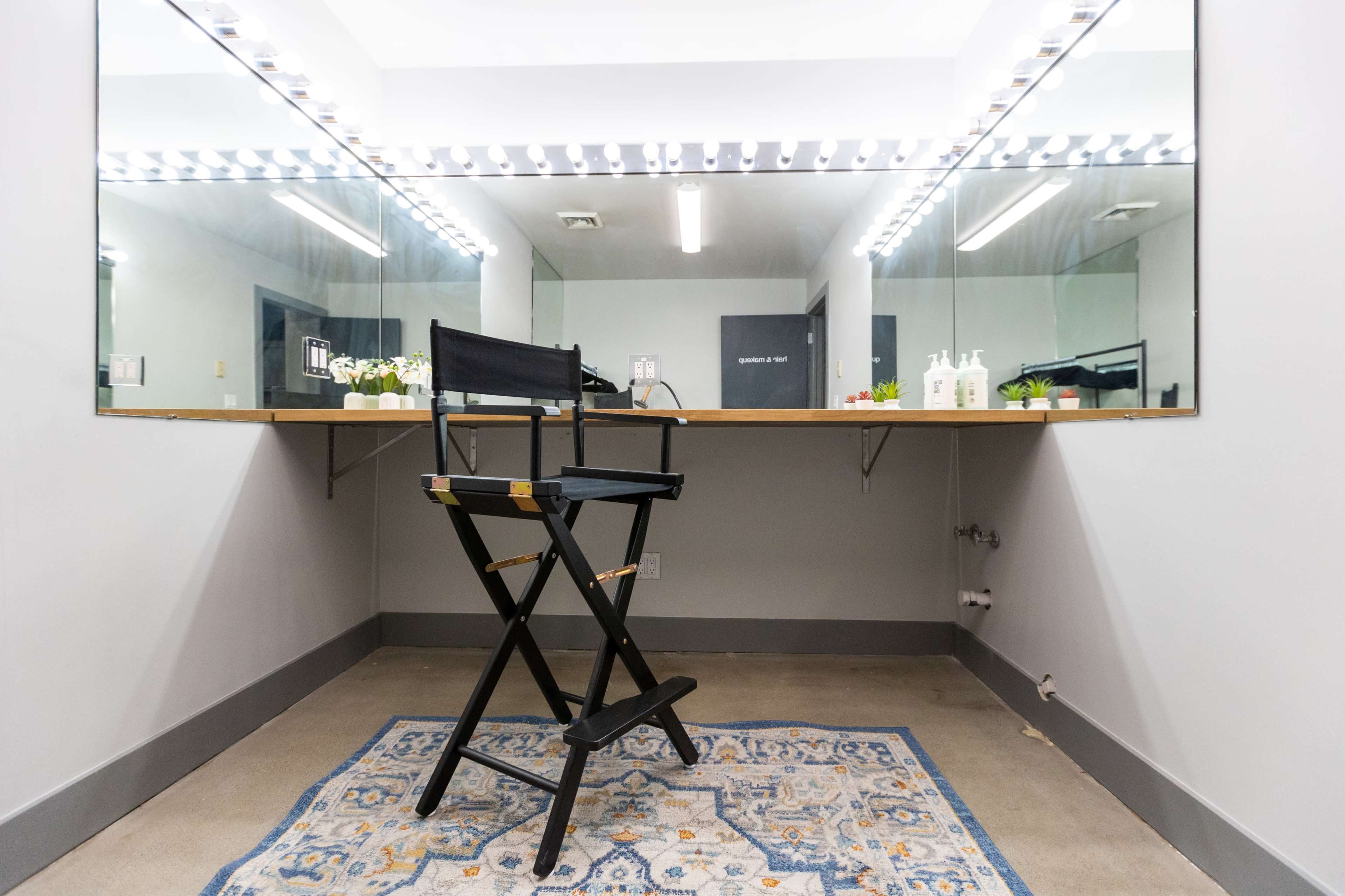 A makeup room features a large mirror with bright lights, a wooden countertop, and a director's chair set on a patterned rug.