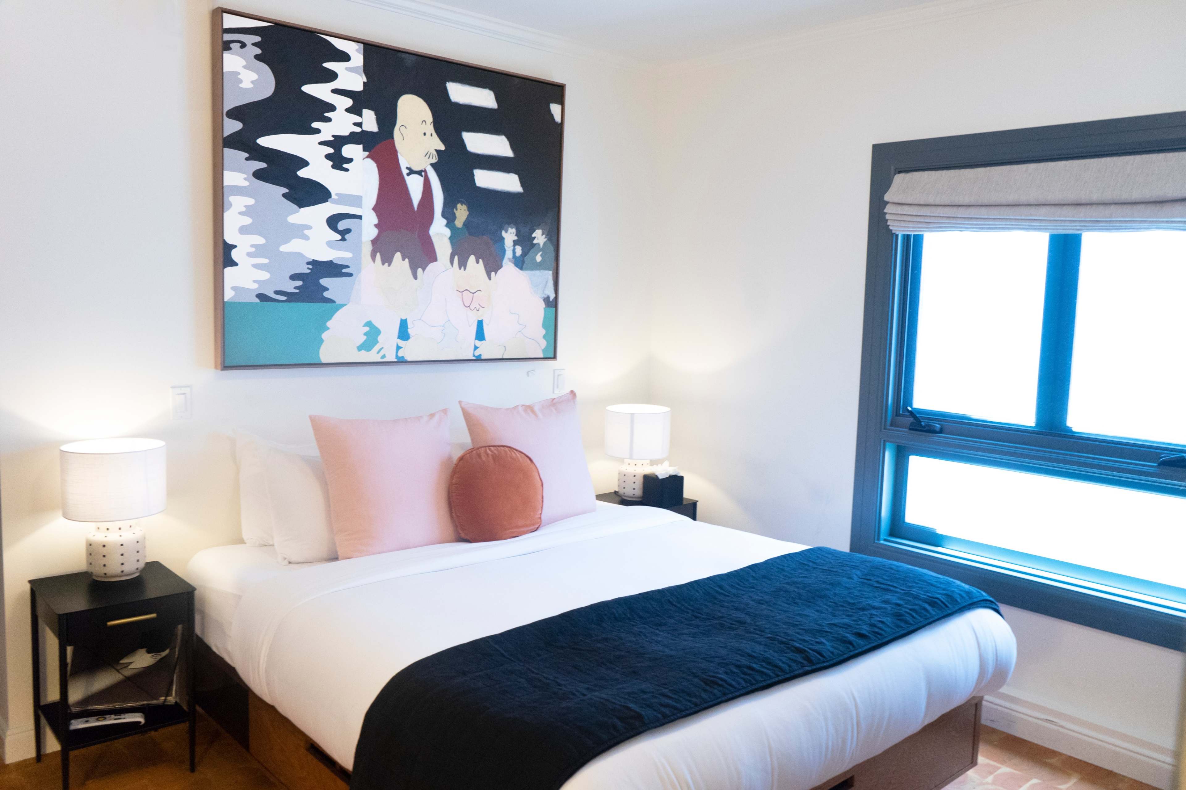 A cozy bedroom featuring a large bed with decorative pillows, two bedside lamps, and a colorful artwork above the bed, along with a window on the right side.