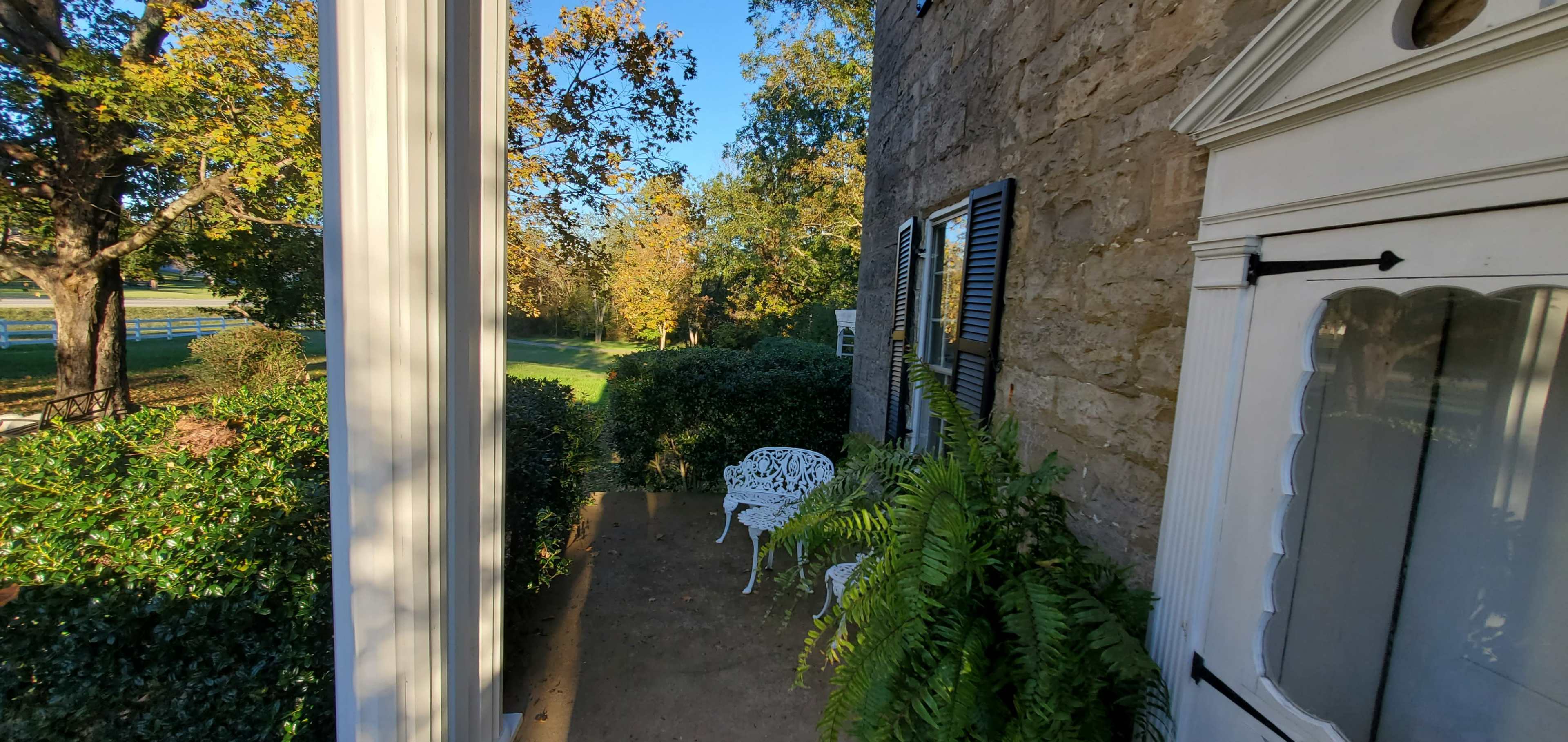 A porch area with a white wrought iron bench set against a stone wall surrounded by greenery.