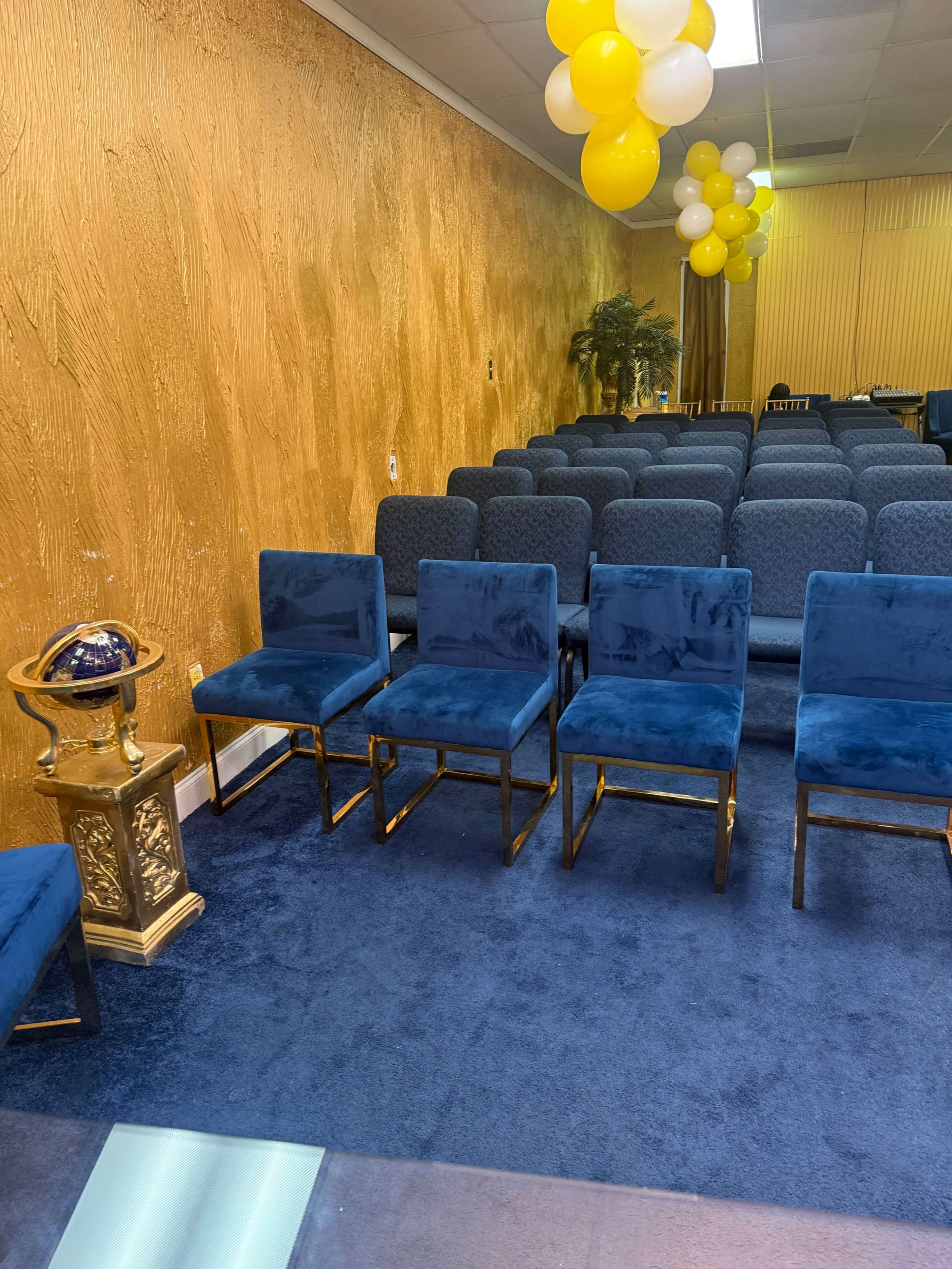 The image shows a seating area in a room with blue velvet chairs arranged in rows, a gold decorative stand with a globe, and yellow and white balloons hanging overhead.