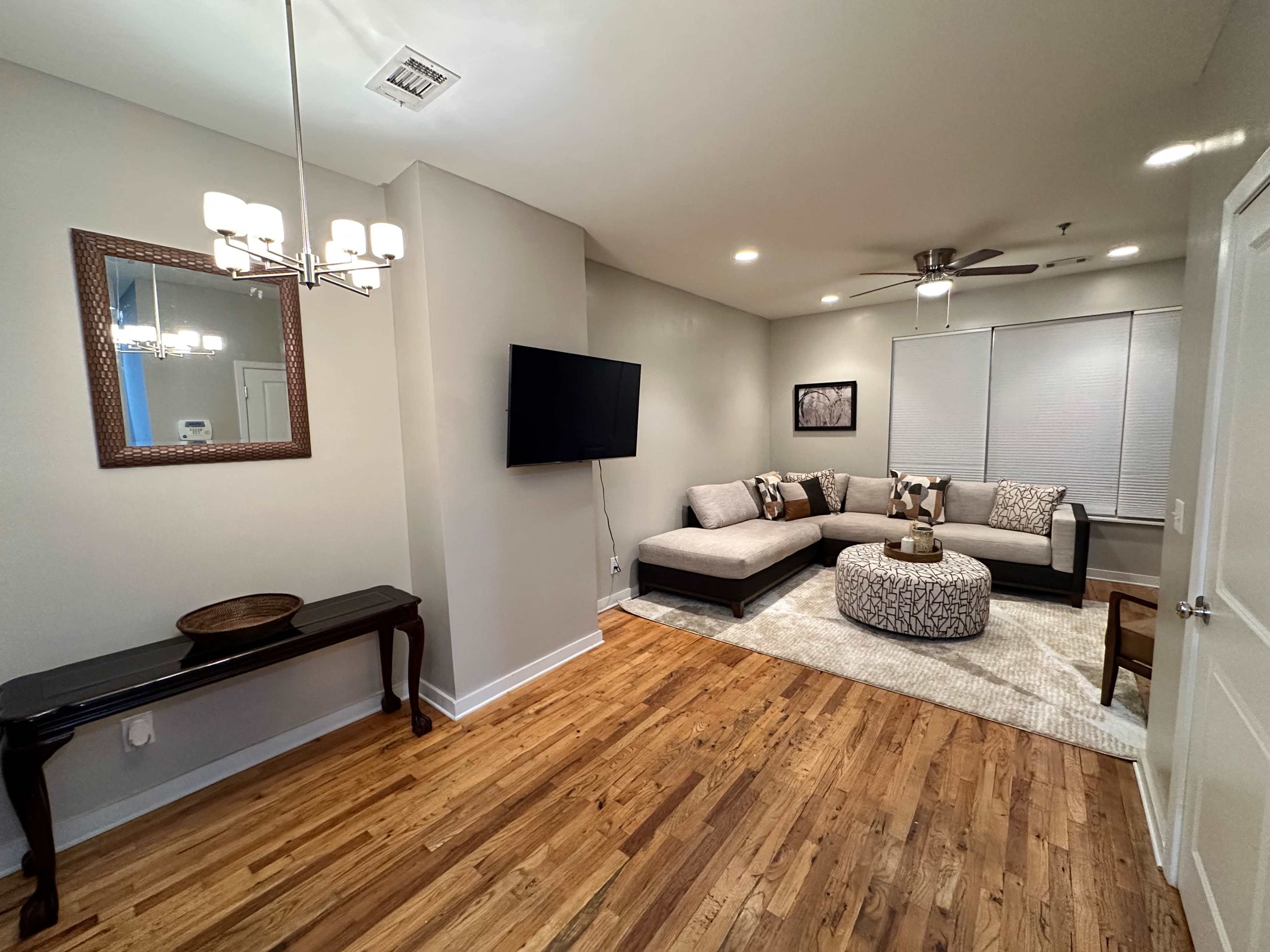 A living room featuring a sectional sofa, a coffee table with decorative items, a wall-mounted television, and wooden flooring.