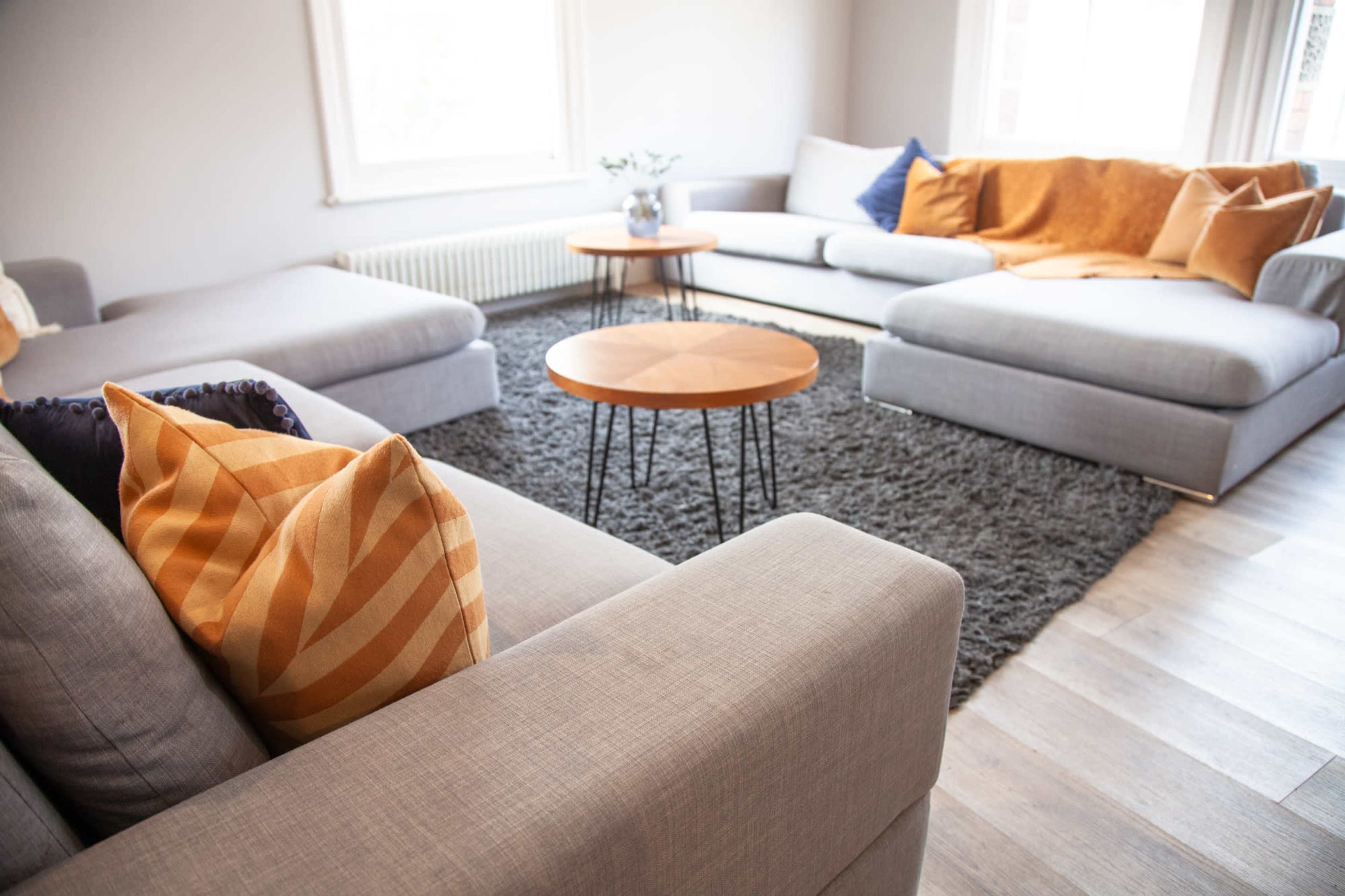 The image shows a modern living room with gray sectional sofas, a wooden coffee table, and decorative pillows arranged on a plush area rug.