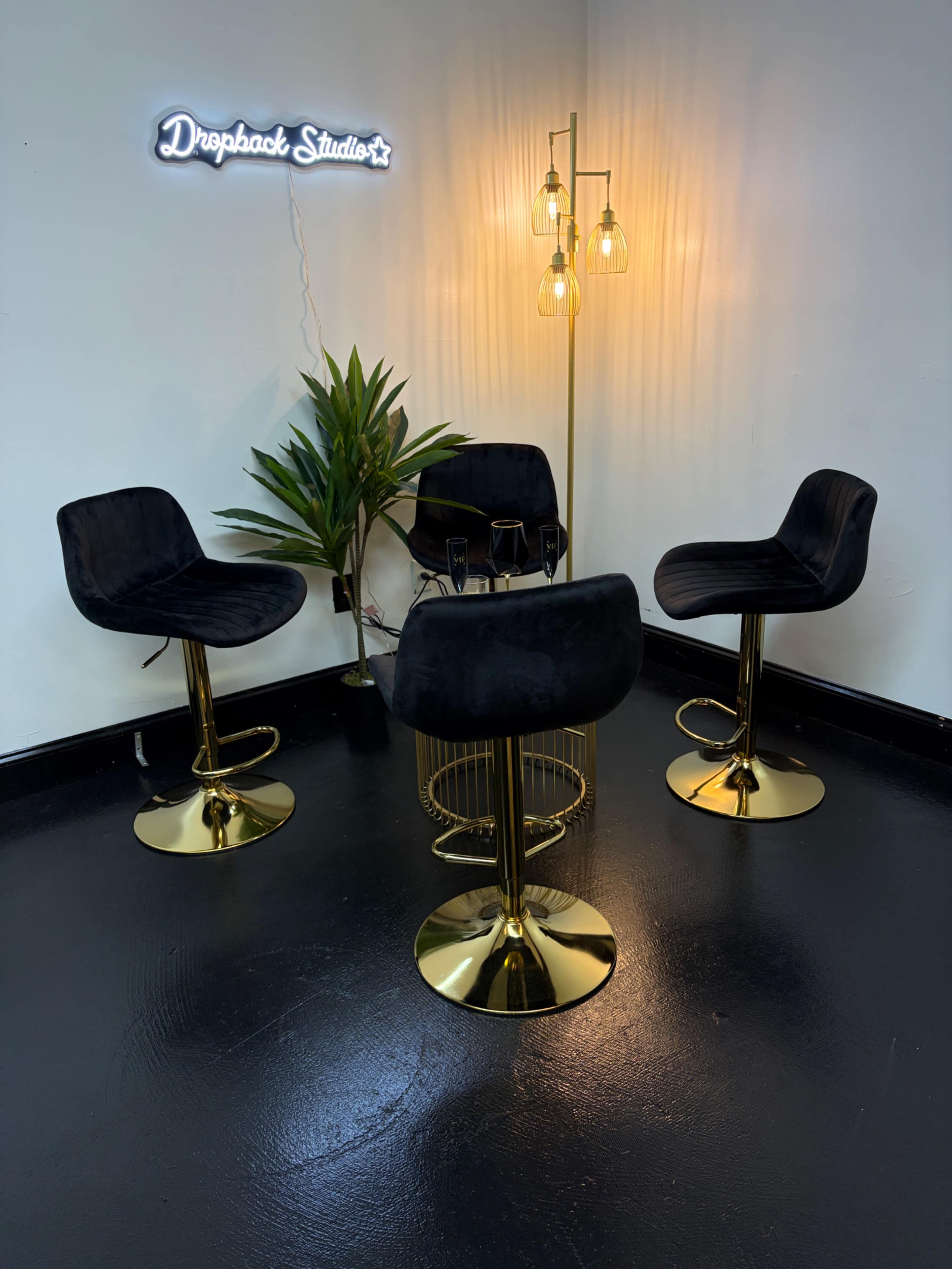 The image shows a stylish studio space with four black bar stools around a small gold table, featuring a modern light fixture and a decorative plant.