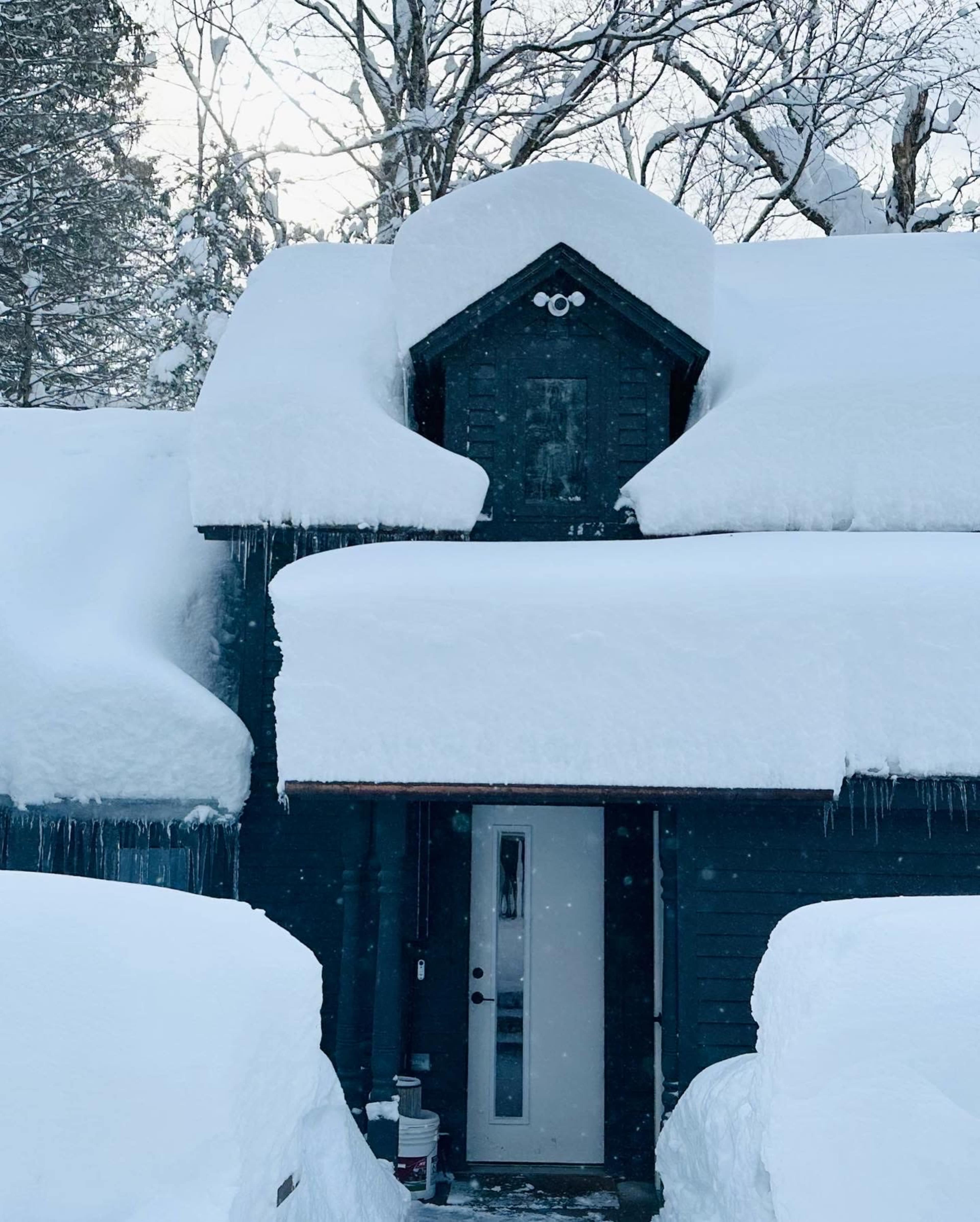 A dark-colored house is covered in thick layers of snow, with snow piled high on the roof and around the entrance.