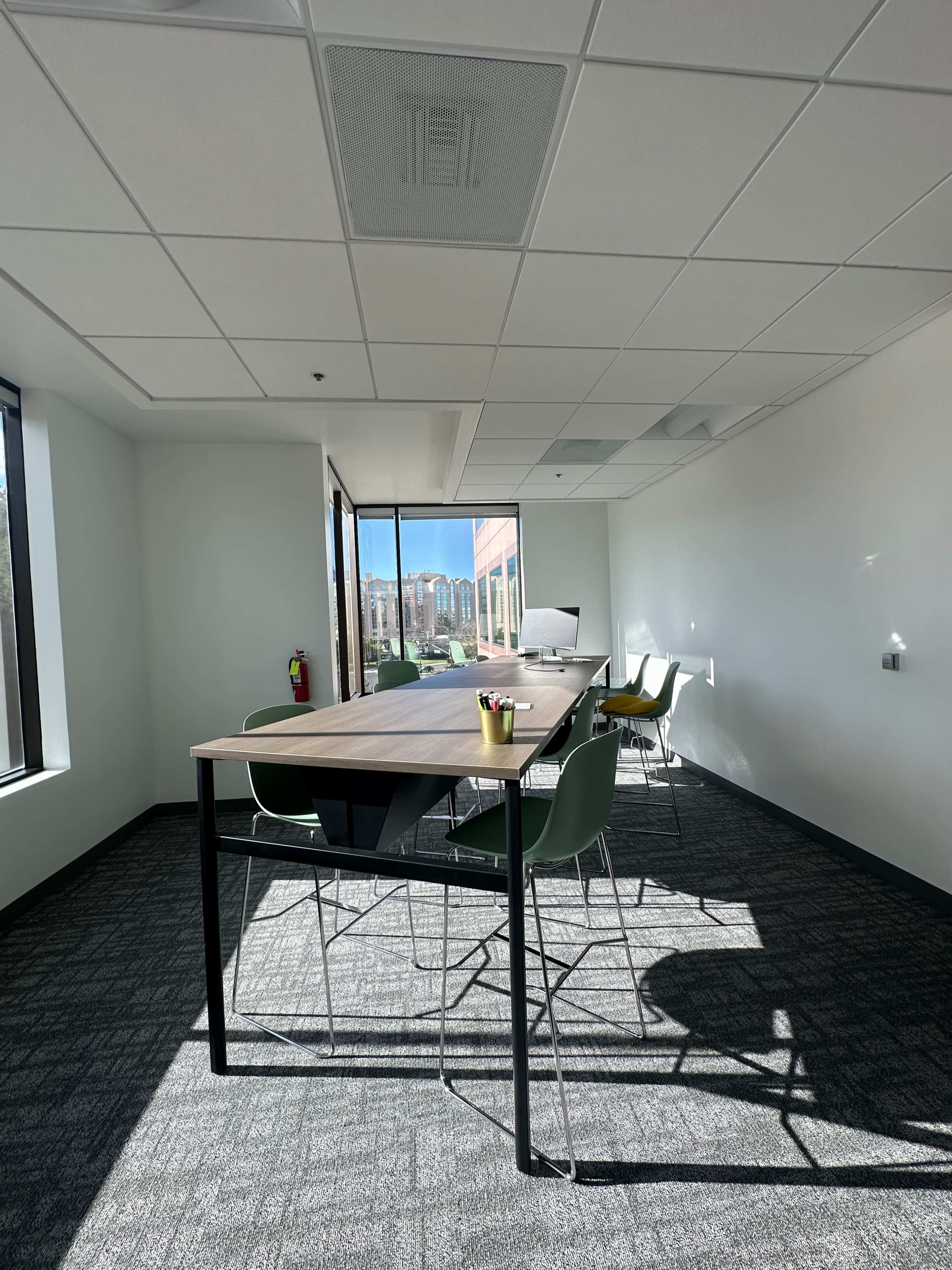 The image shows a bright, modern office conference room featuring a large table surrounded by green chairs and a view of a cityscape through the windows.