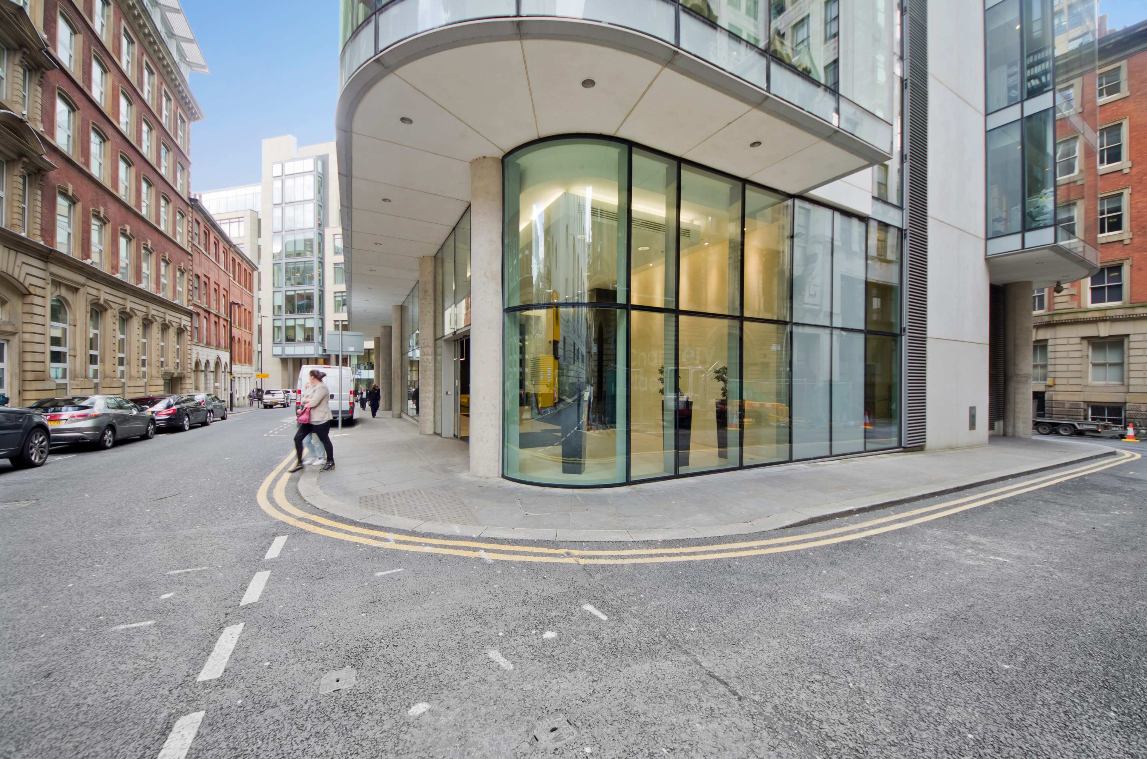 A modern glass-fronted building intersects with a narrow street lined with parked cars and pedestrians.