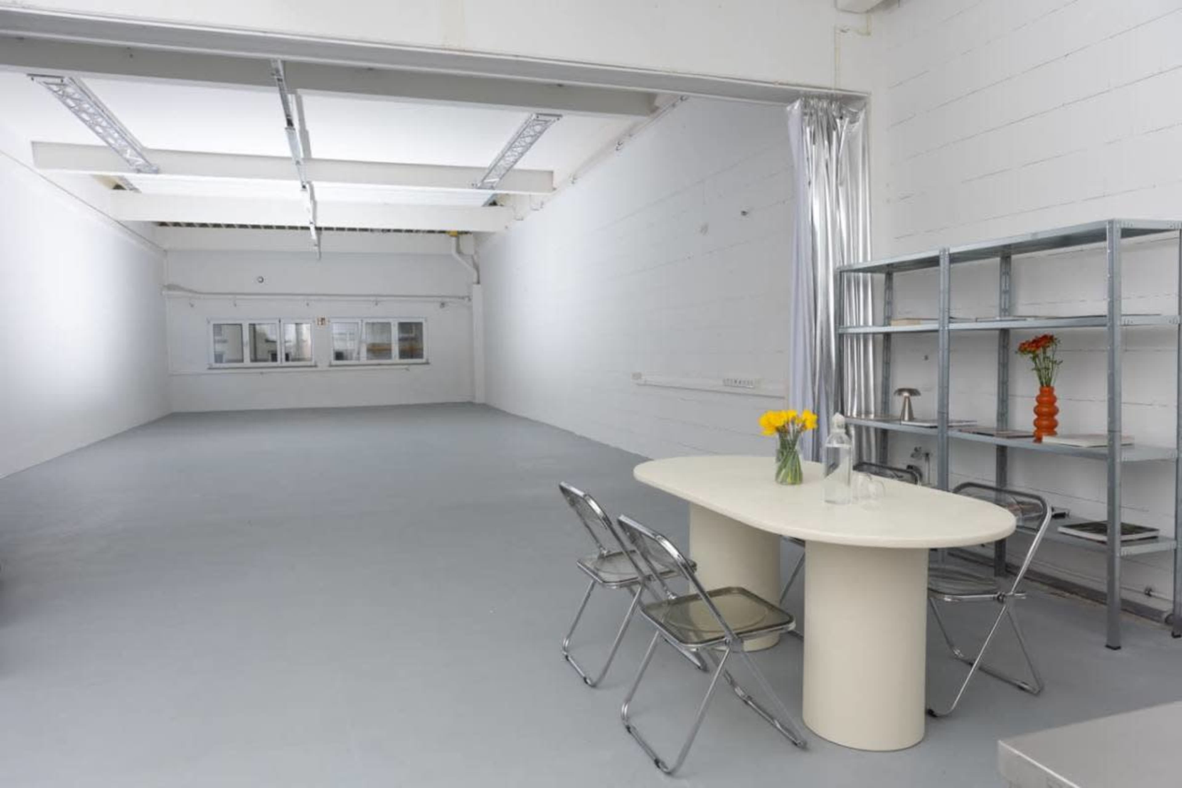 The image shows a minimalistic, empty studio space with a round table and chairs arranged near a metal shelf.