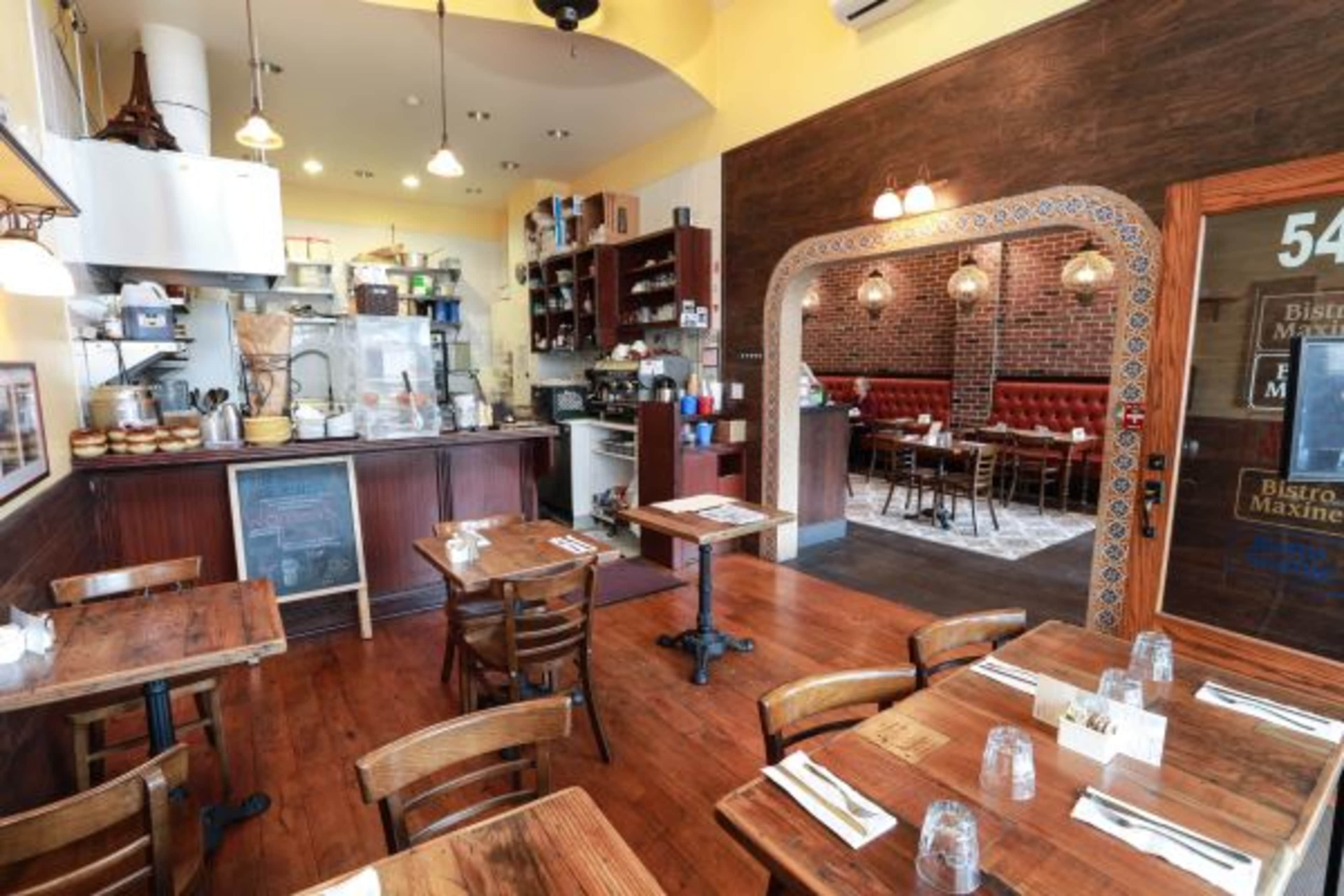 A cozy restaurant interior features wooden tables and chairs, a structured counter with kitchen equipment, and a brick accent wall in the dining area.