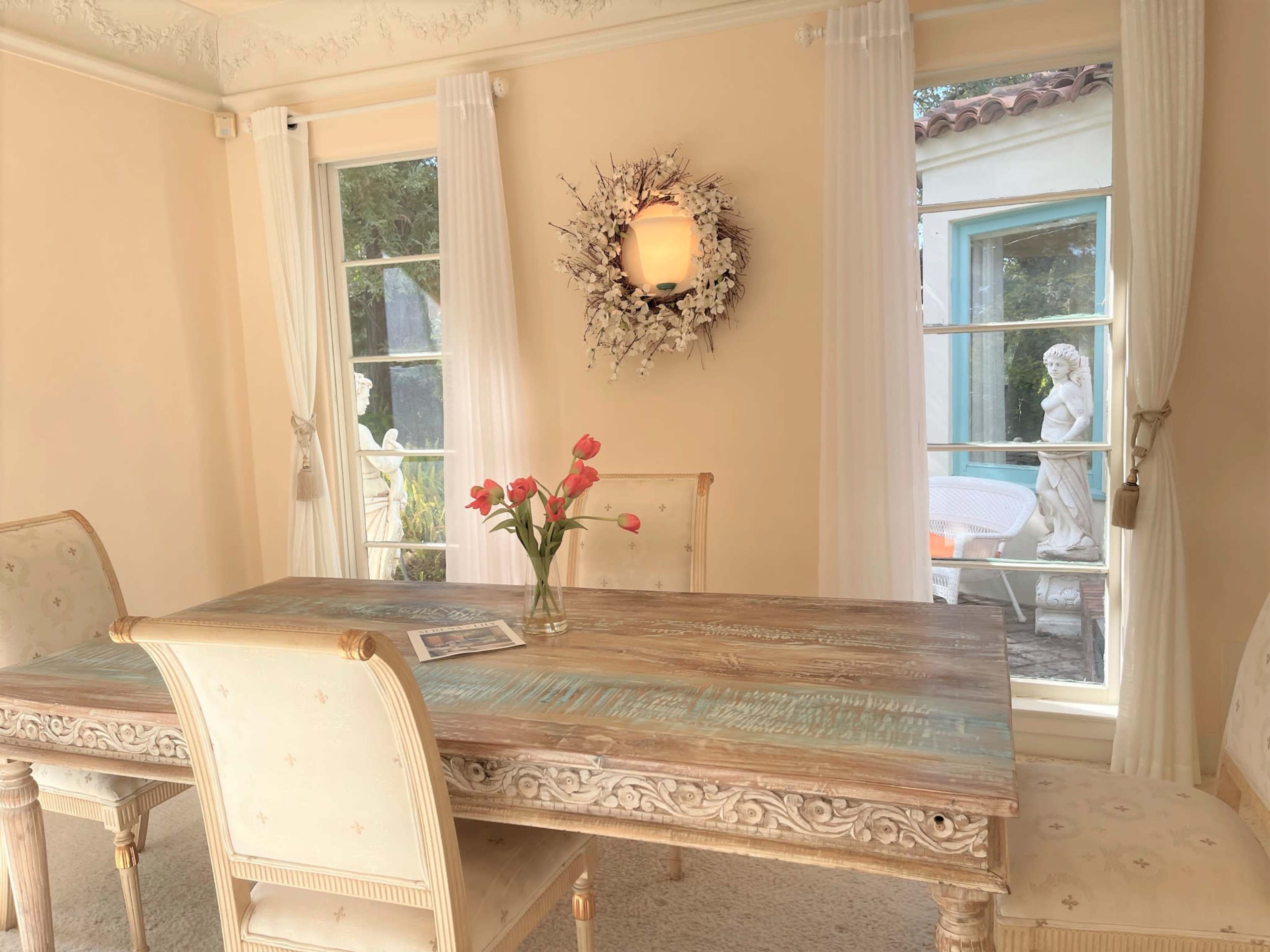 A wooden dining table with a floral centerpiece is set in a room featuring large windows, white drapes, and a wall-mounted light fixture.