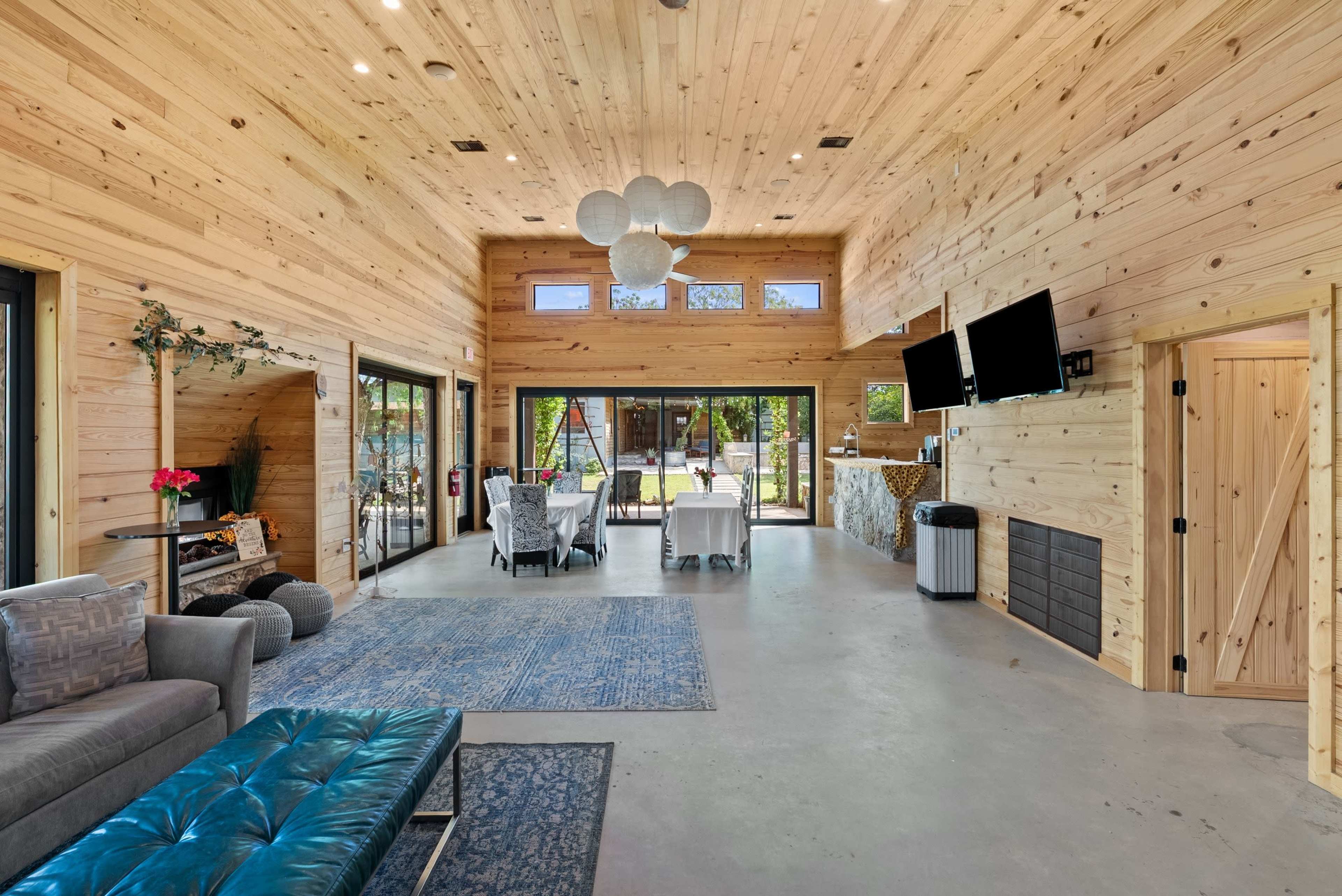 The image shows a spacious room with wooden walls and ceilings, featuring a seating area, dining setup, and large glass doors opening to an outdoor space.