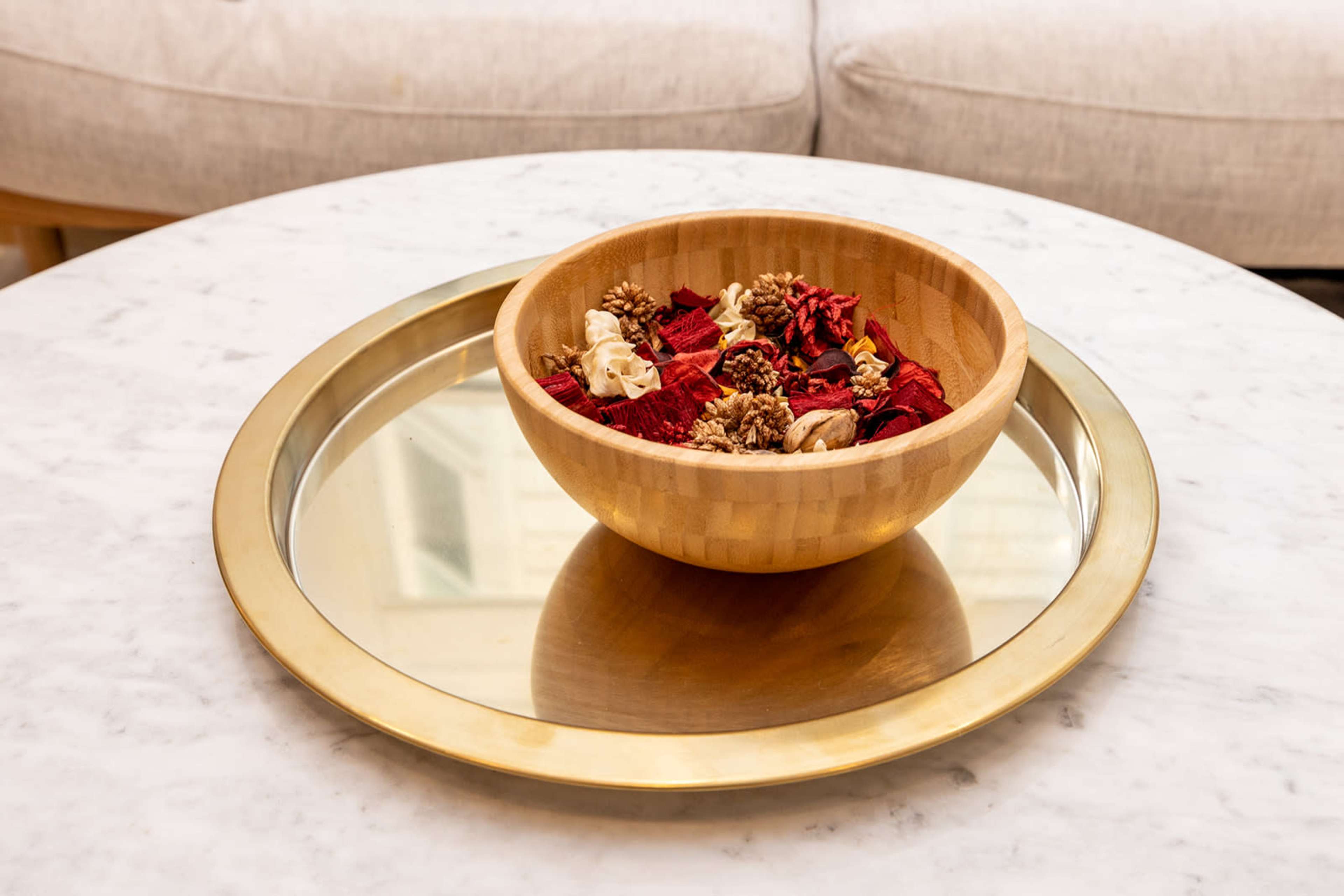 A wooden bowl filled with decorative items rests on a round gold tray placed on a marble table.