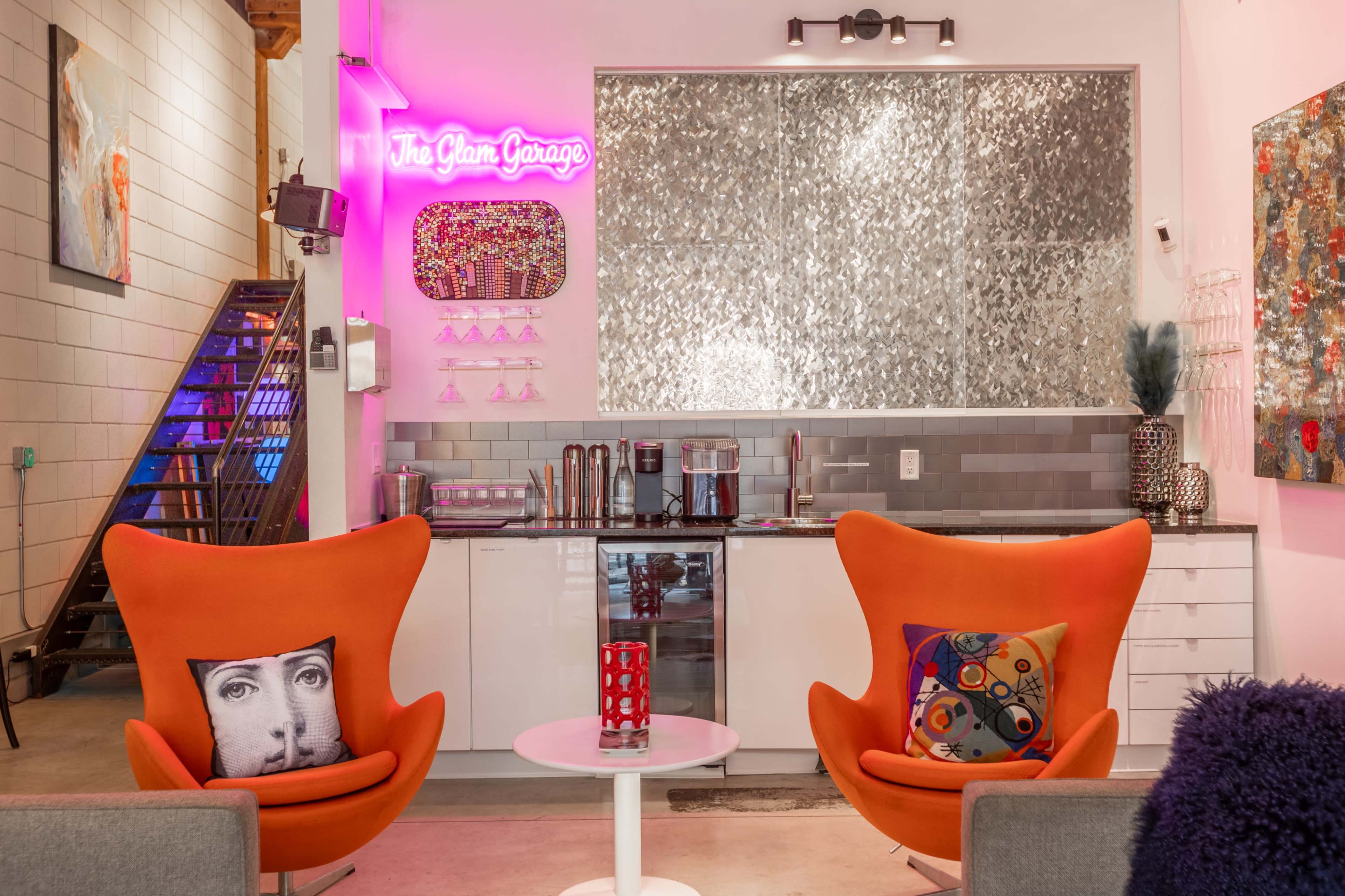 The image shows a modern space featuring two vibrant orange chairs facing a small white table, with a stylish bar area in the background adorned with a decorative neon sign and various art pieces.