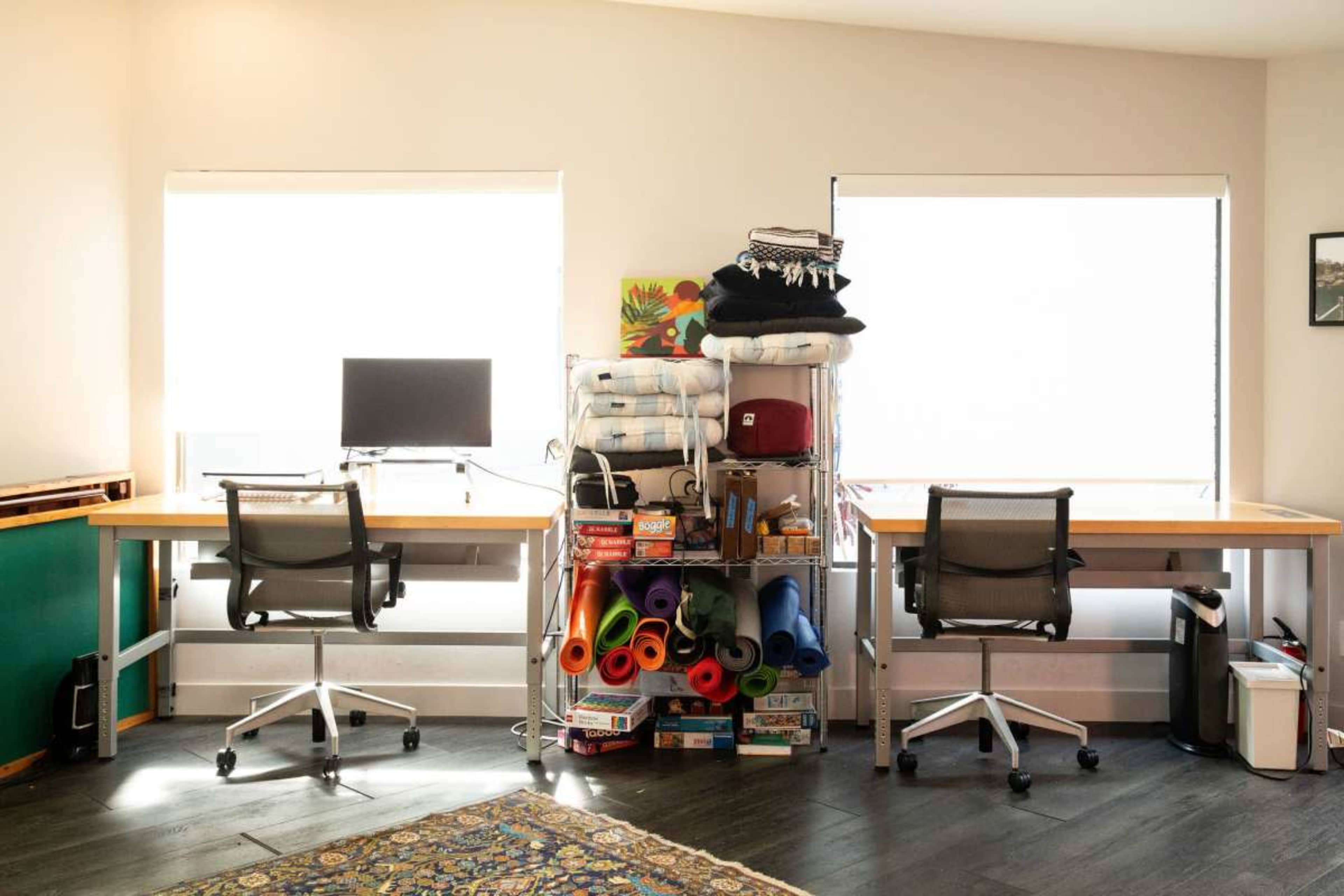 The image shows a bright room with two desks, each equipped with a chair and a monitor, and a shelving unit stacked with various items next to the windows.