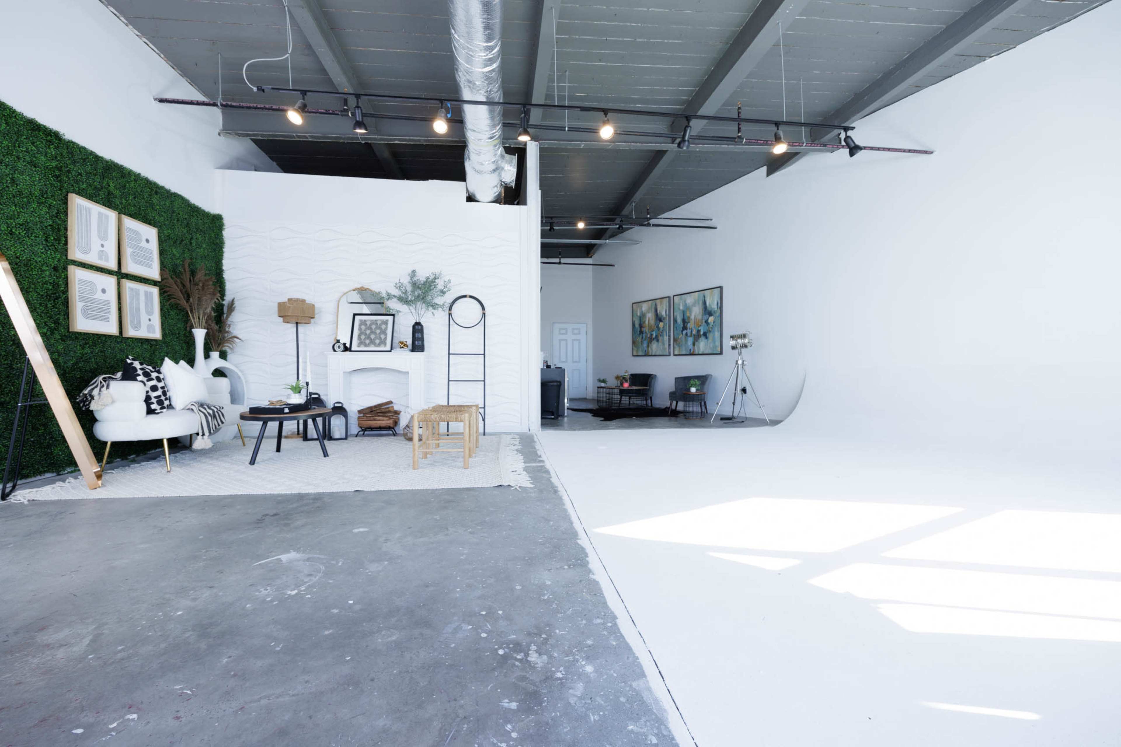The image shows a spacious, bright studio interior with a seating area on one side and an open space on the other, featuring minimalist décor and large windows.
