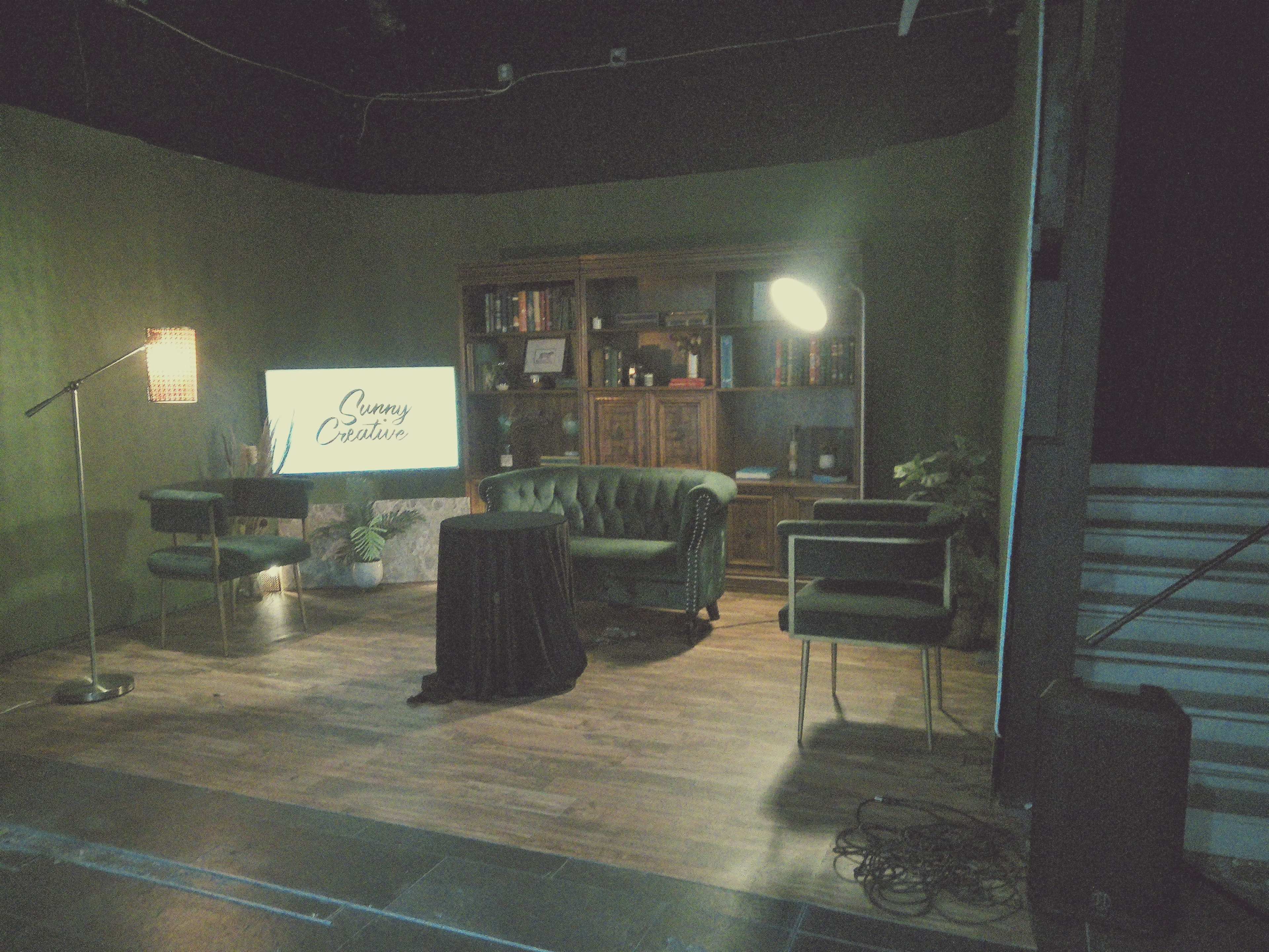 The image shows a studio setup with a green wall, a wooden bookcase, a round black table, two green chairs, and a floor lamp, all arranged around a television displaying the words "Sunny Creative."