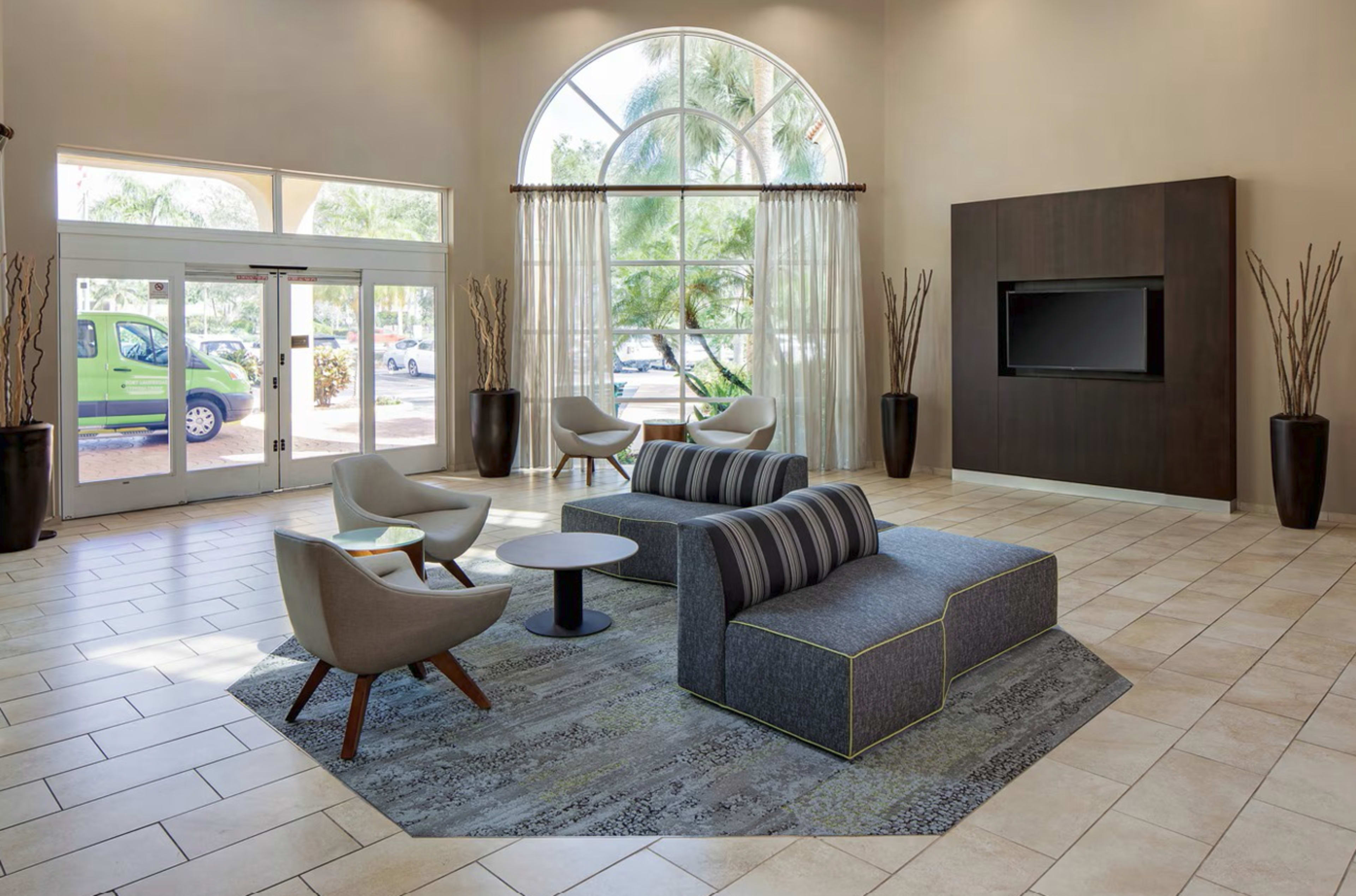 The image shows a modern lobby area featuring a seating arrangement with gray furniture, a coffee table, and large windows allowing natural light to enter.