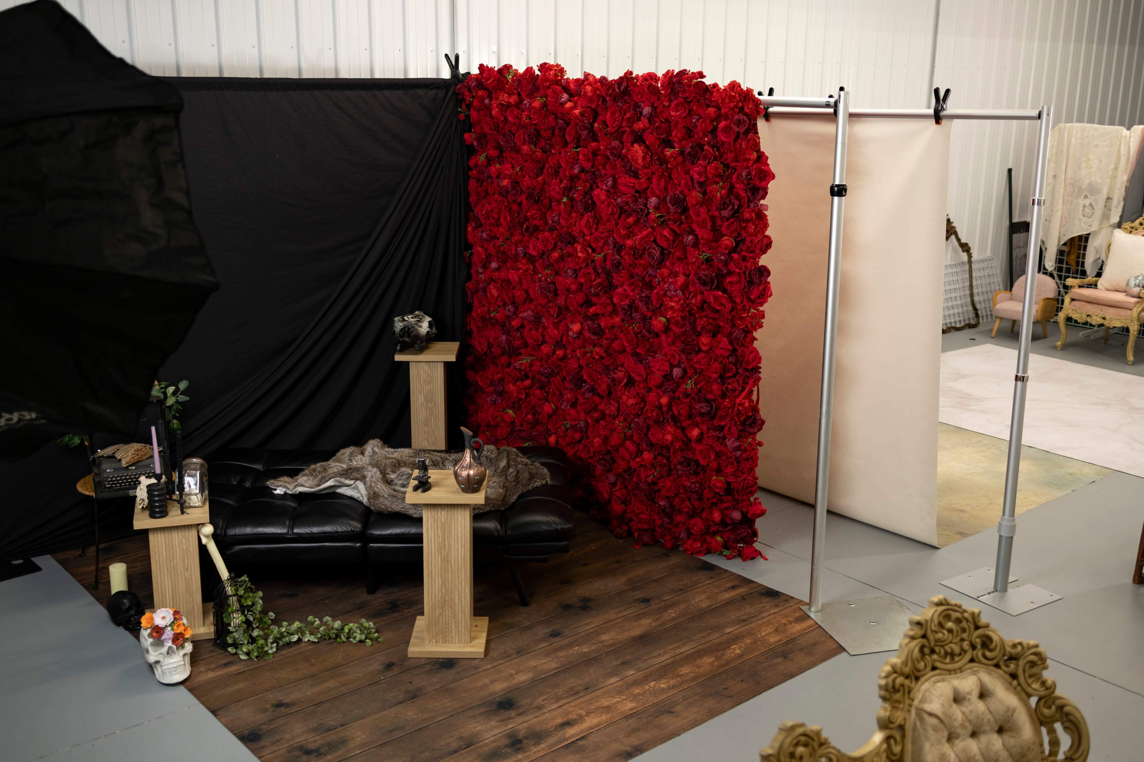 The image shows a photography studio setup featuring a black backdrop, a vibrant red floral wall, a black leather sofa, and various props arranged on wooden tables.