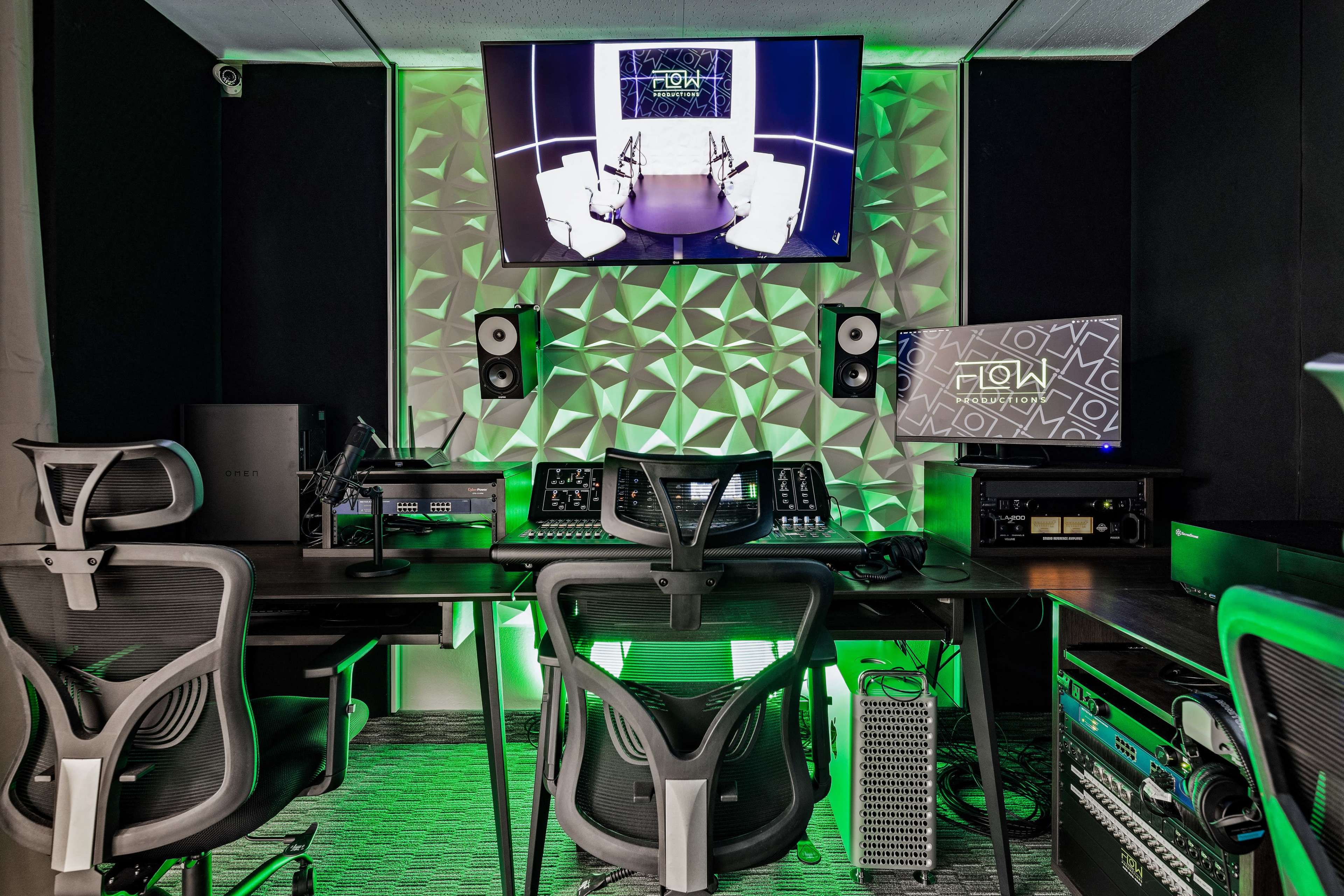 A modern audio-visual control room with multiple computer monitors, a sound mixing console, and three office chairs arranged around a large table.