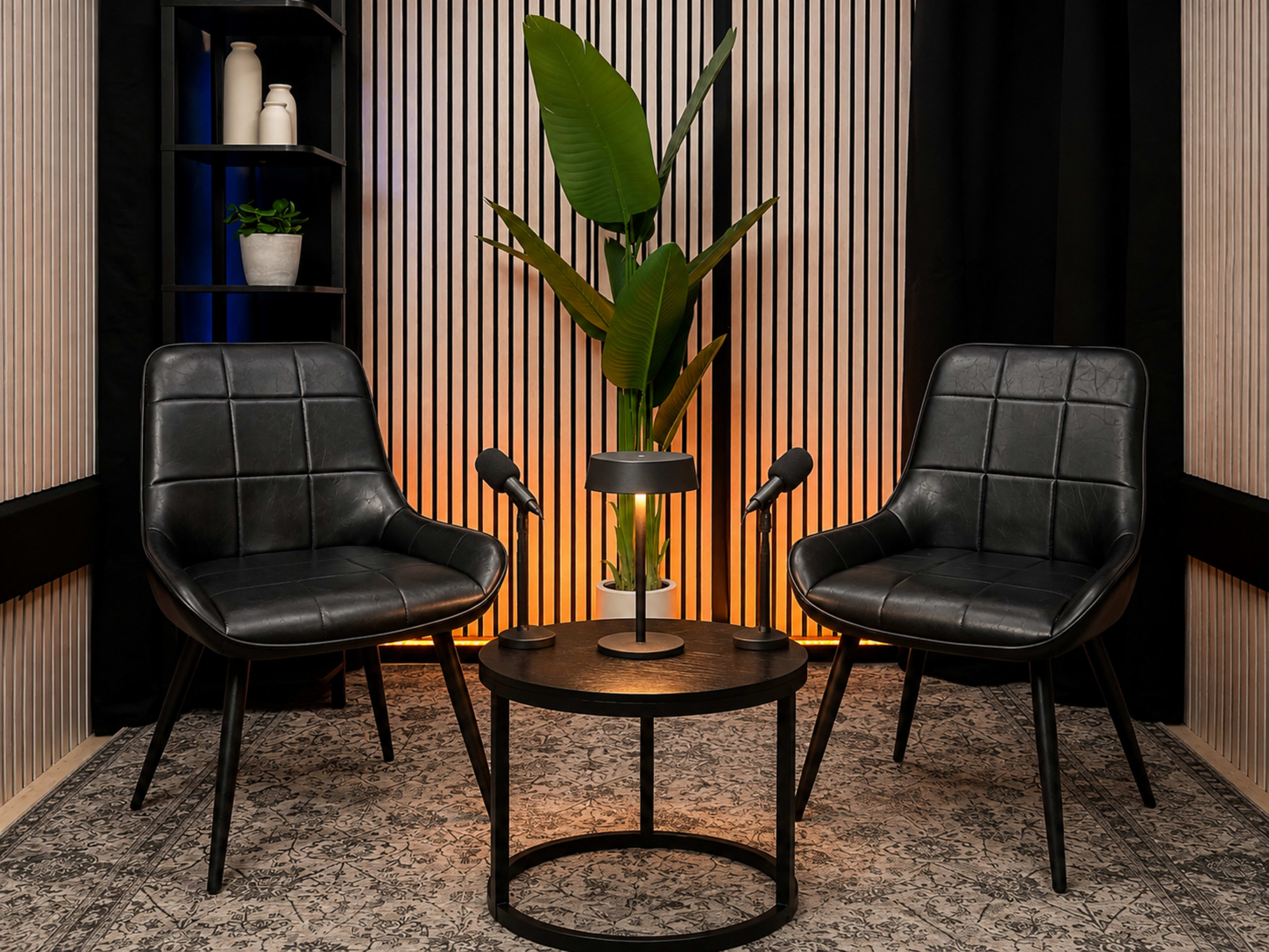The image shows a modern seating arrangement with two black chairs facing a circular table, featuring a lamp and a potted plant, against a backdrop of striped walls and ambient lighting.