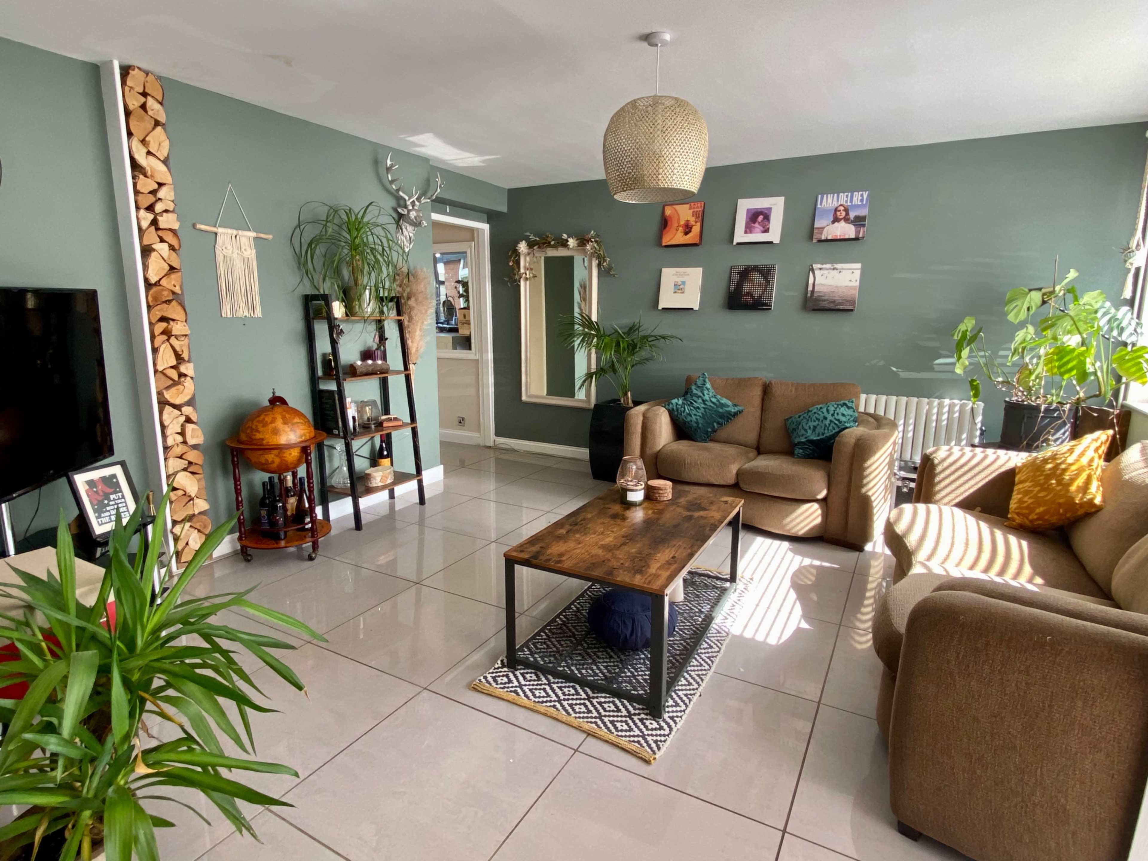 A living room with two brown sofas, a wooden coffee table, a bookshelf, and several plants, featuring light-colored tiled flooring and a green wall.