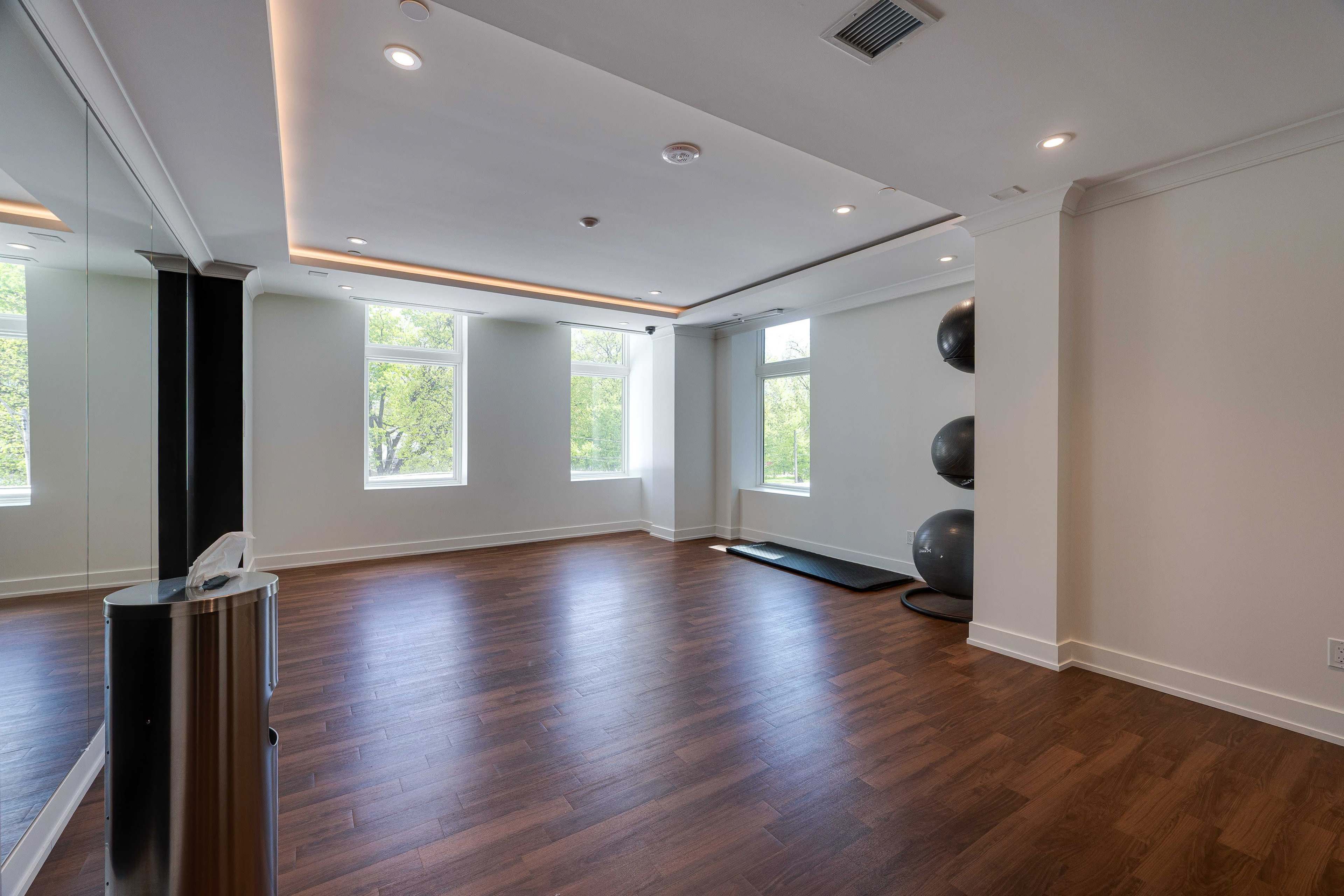 The image shows a spacious, well-lit room with hardwood floors, large windows, and a small area for exercise equipment, including three stability balls against the wall.