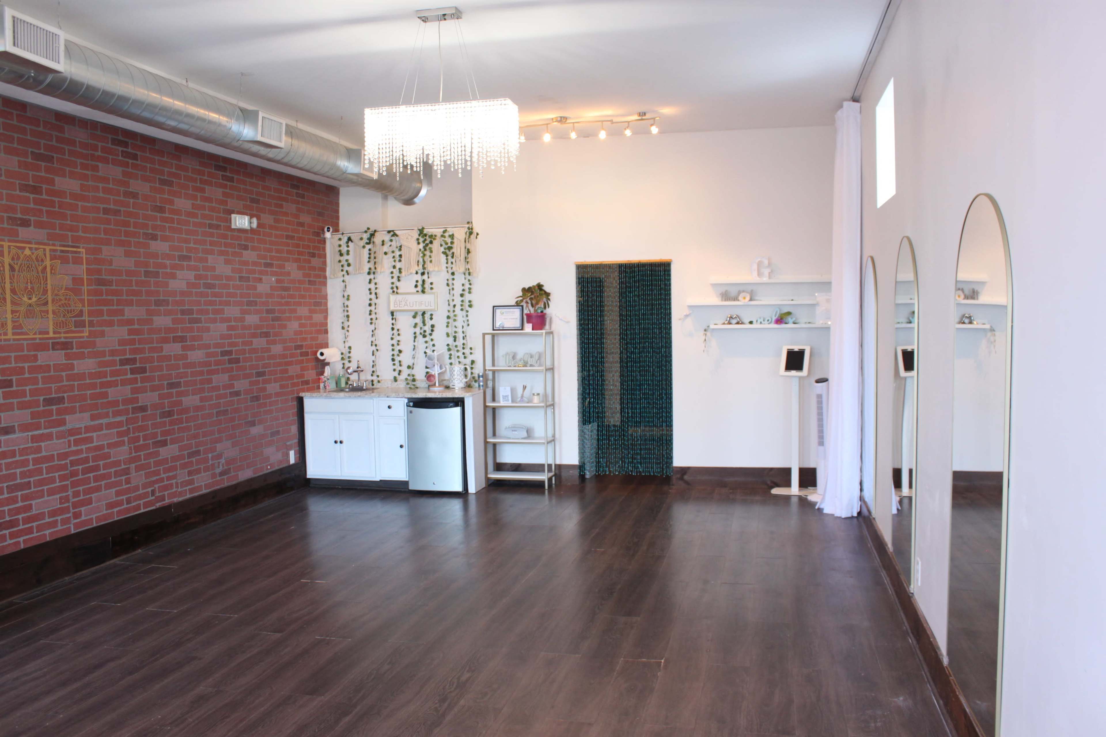 The image shows a spacious room with exposed brick walls, a small kitchenette area, mirrors, and a green tile accent wall.