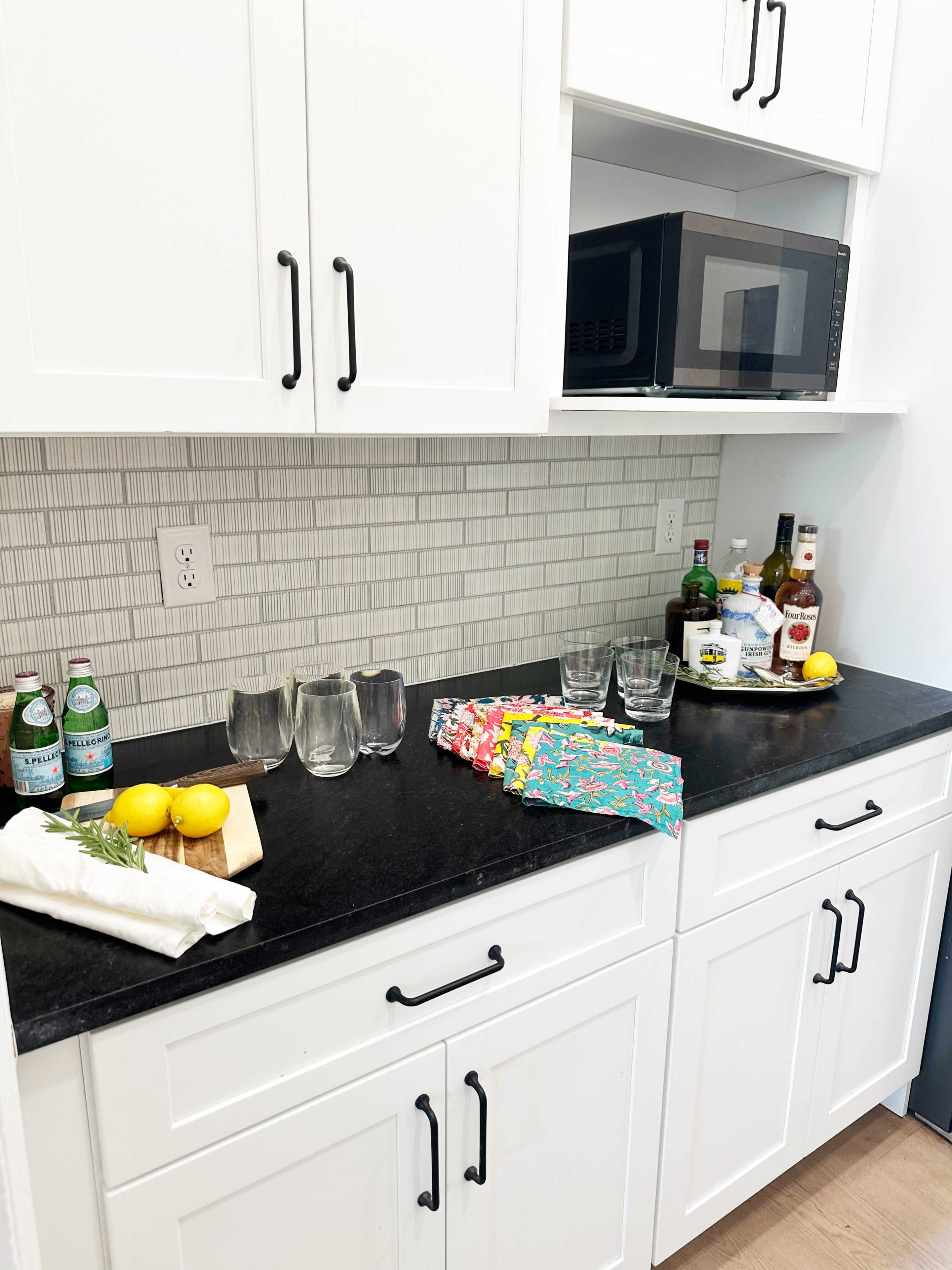 A kitchen countertop features a row of glassware, a cutting board with lemons, a collection of drinks, and a microwave above white cabinets with black handles.