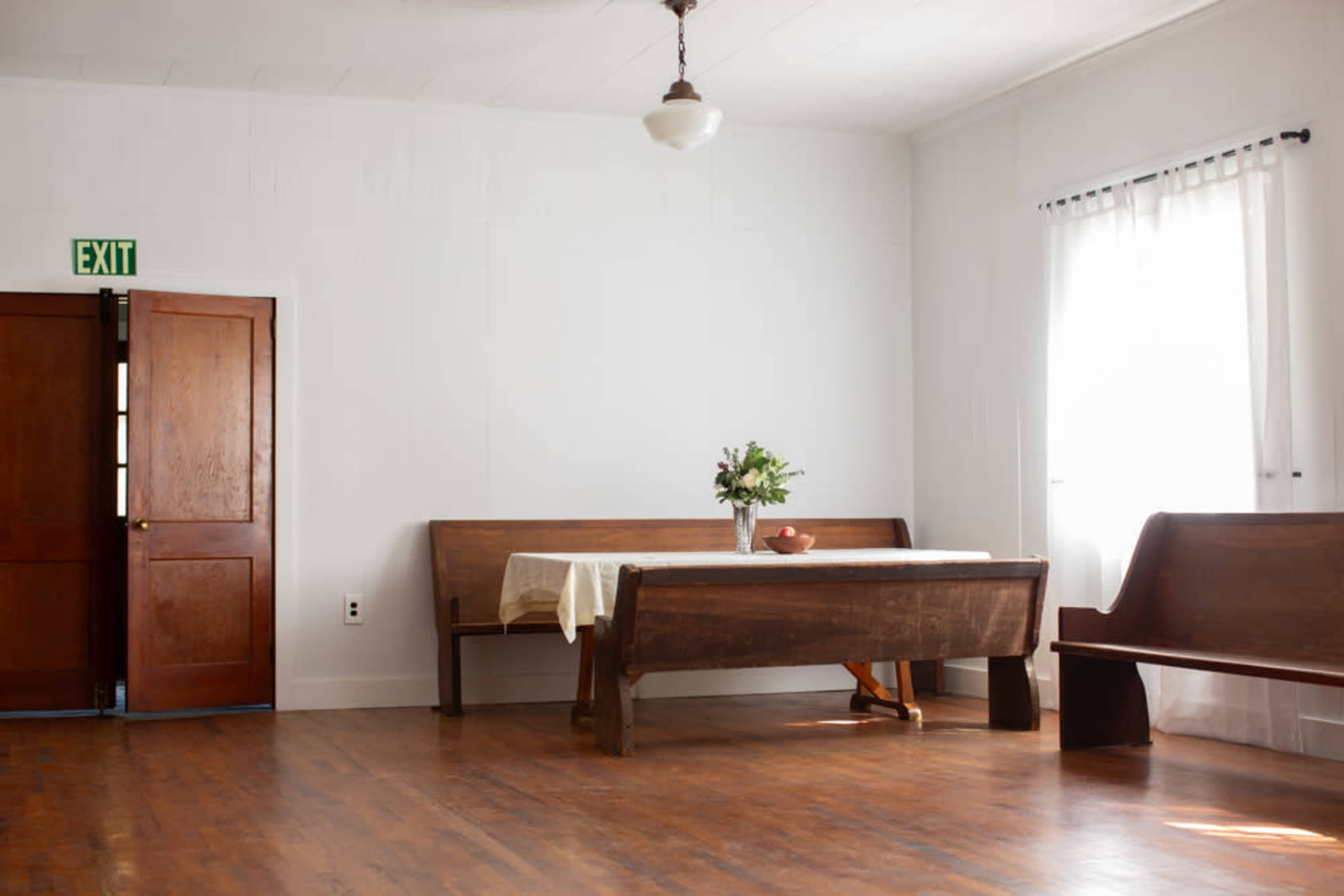 A sparsely furnished room featuring a wooden table with a centerpiece, two long benches, and a door leading outside.