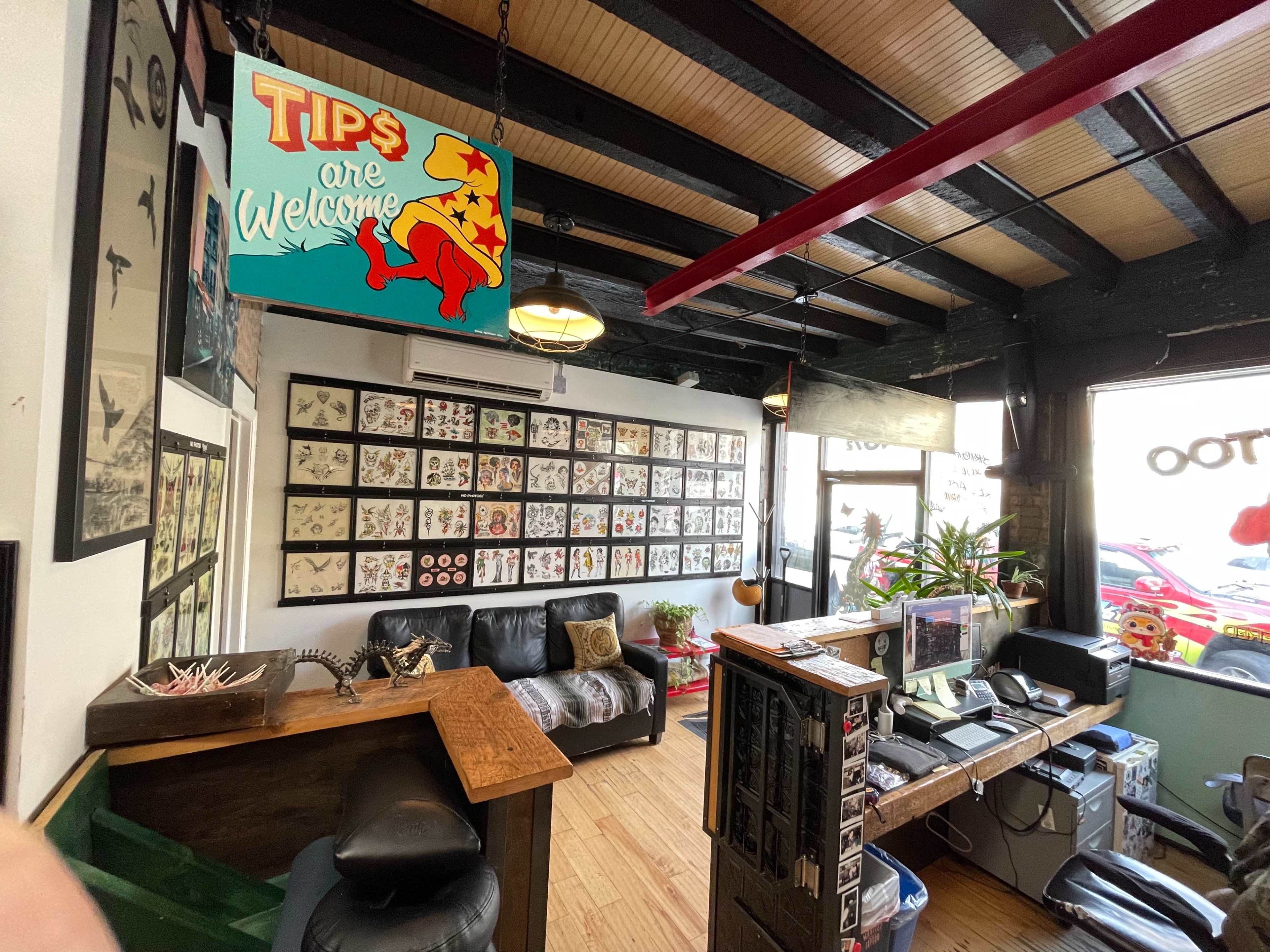 The image shows an interior space of a tattoo studio with a reception area displaying a colorful sign that says "Tips are Welcome," a wall filled with tattoo designs, and a seating area with a couch.