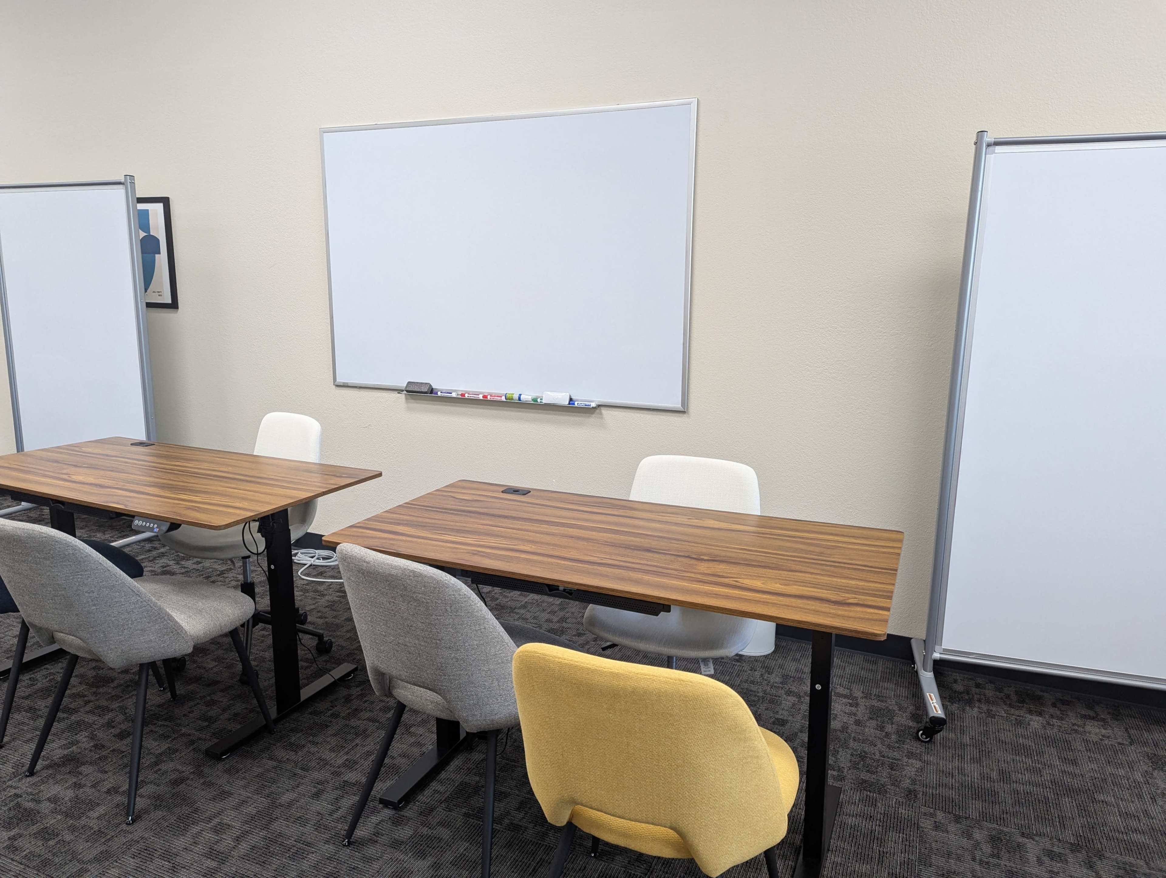 A sparsely furnished meeting room features two wooden tables, several chairs, and a whiteboard mounted on the wall.