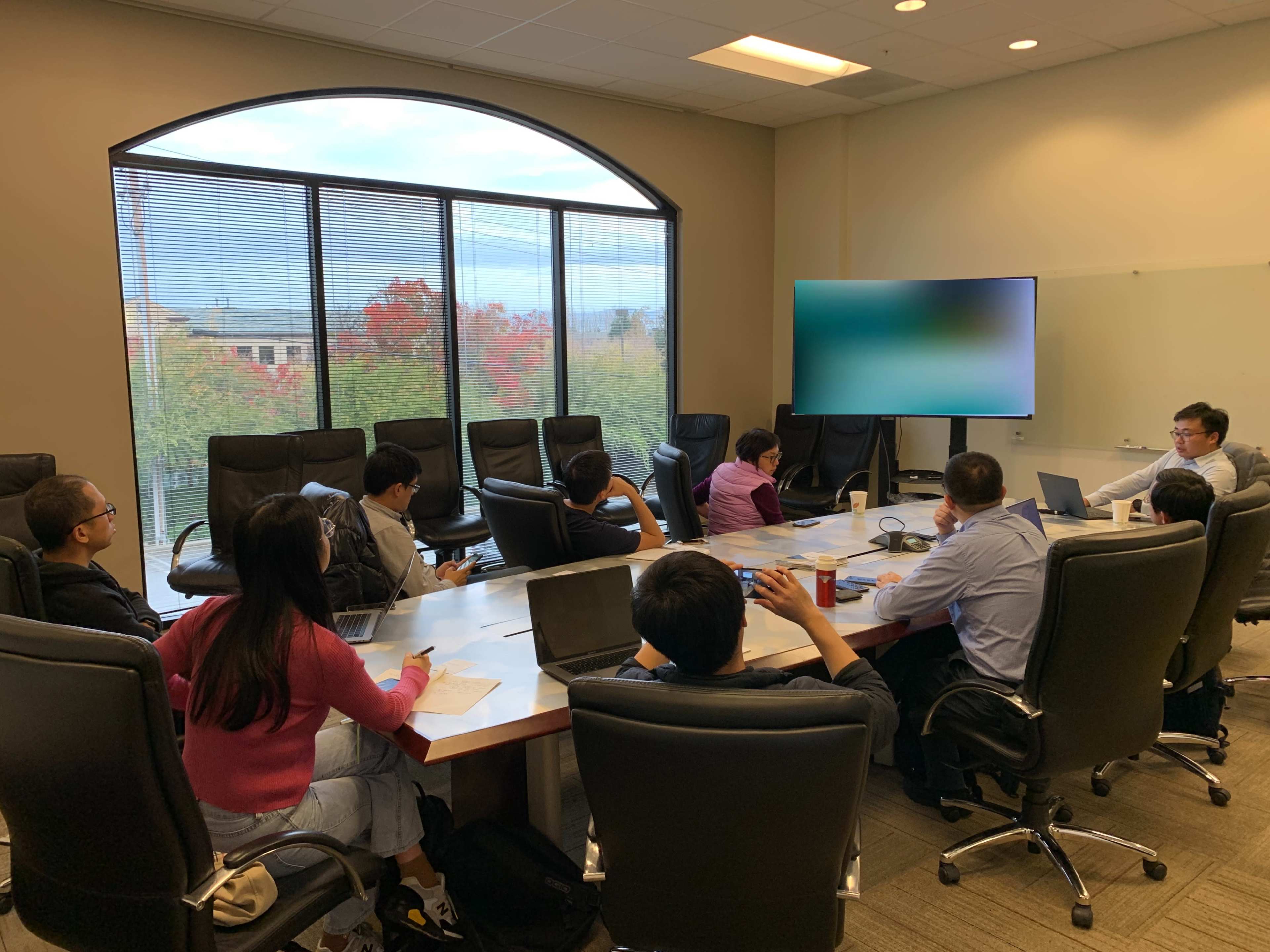 A group of people is seated around a large conference table in a meeting room, with laptops and a large screen displaying a blurred image in the background.