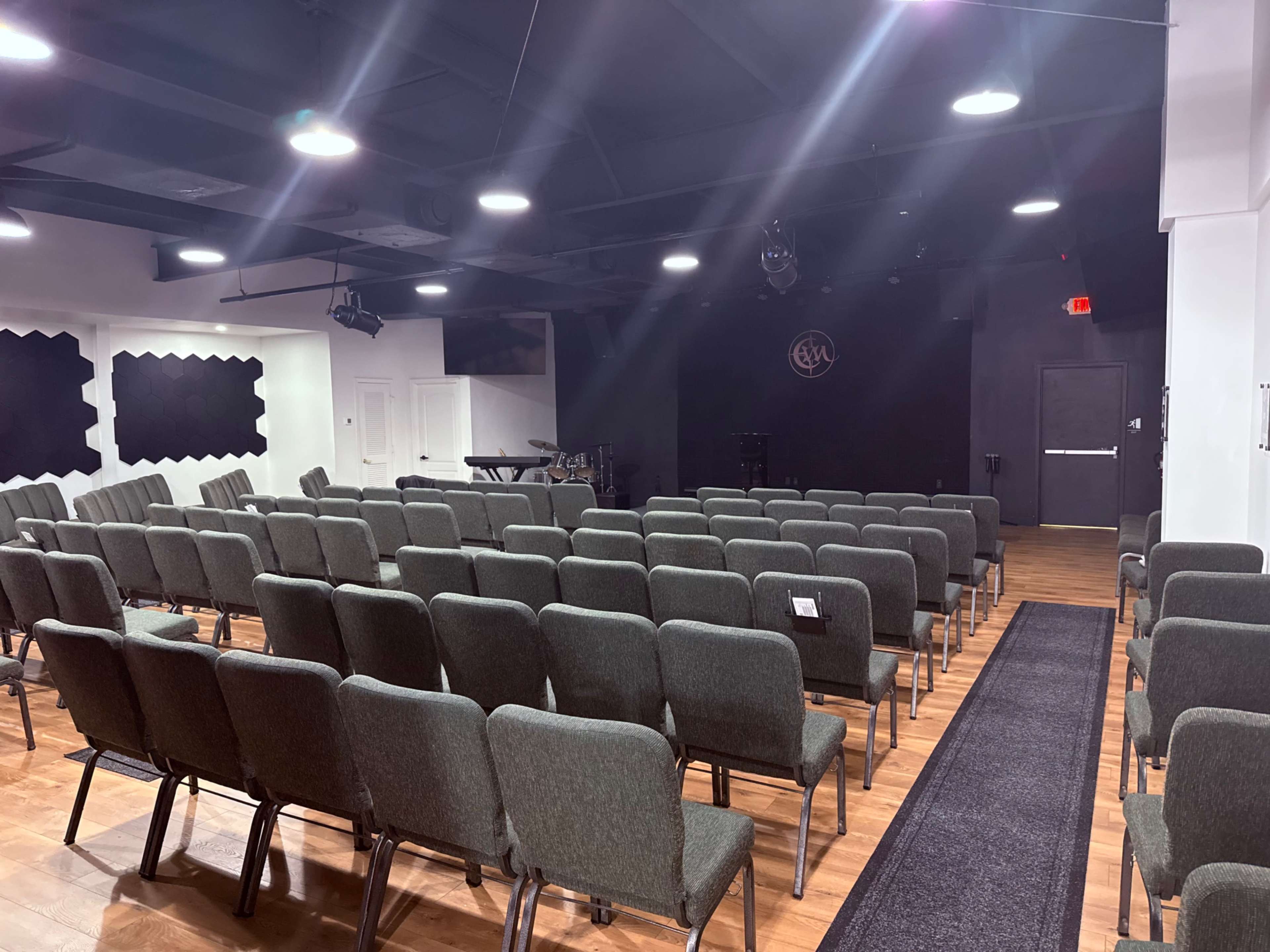 The image shows an indoor auditorium with rows of green chairs facing a black stage, illuminated by overhead lighting.