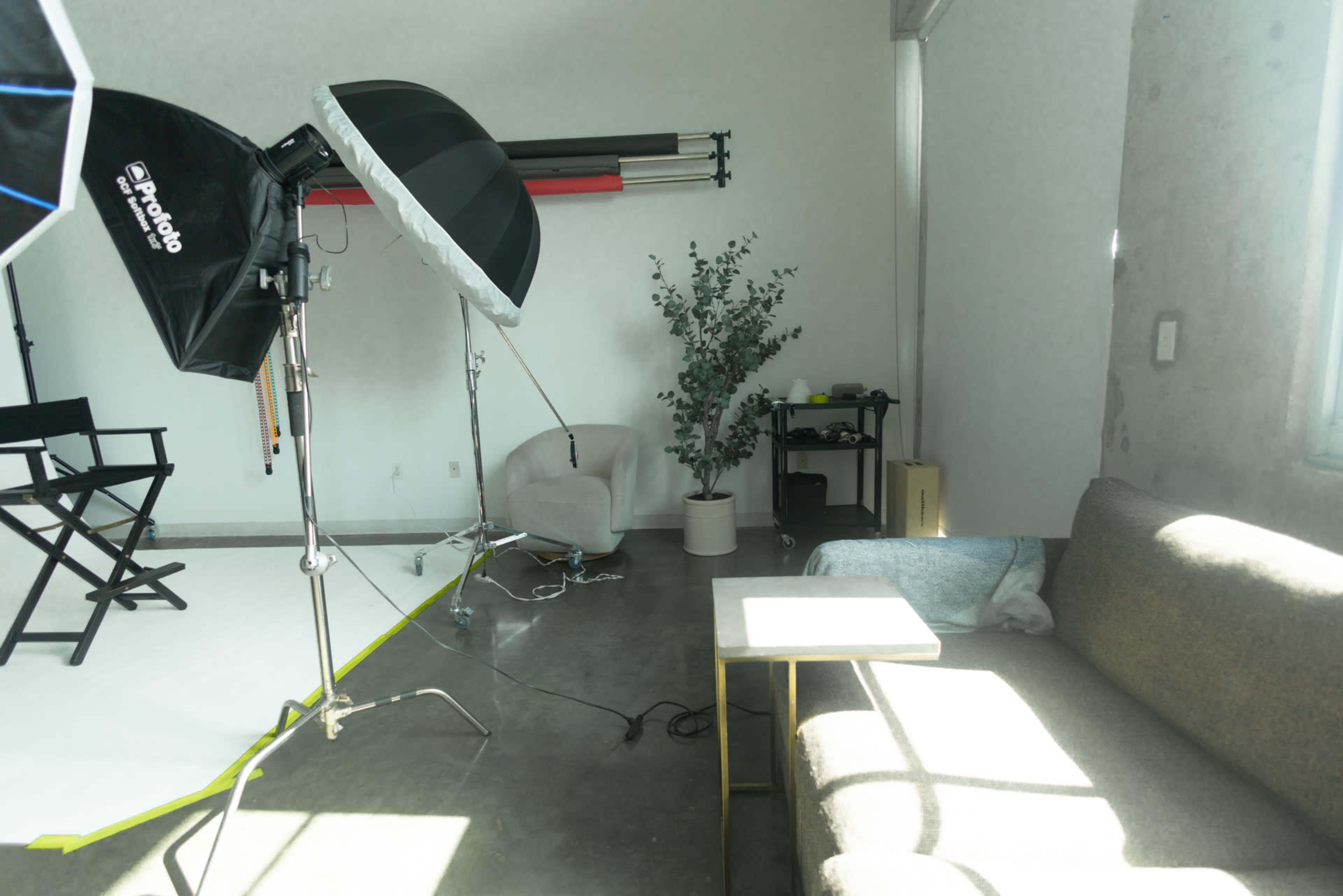 The image shows a photography studio setup with a large umbrella light, a director's chair, a couch, a small table, and a potted plant in the corner.