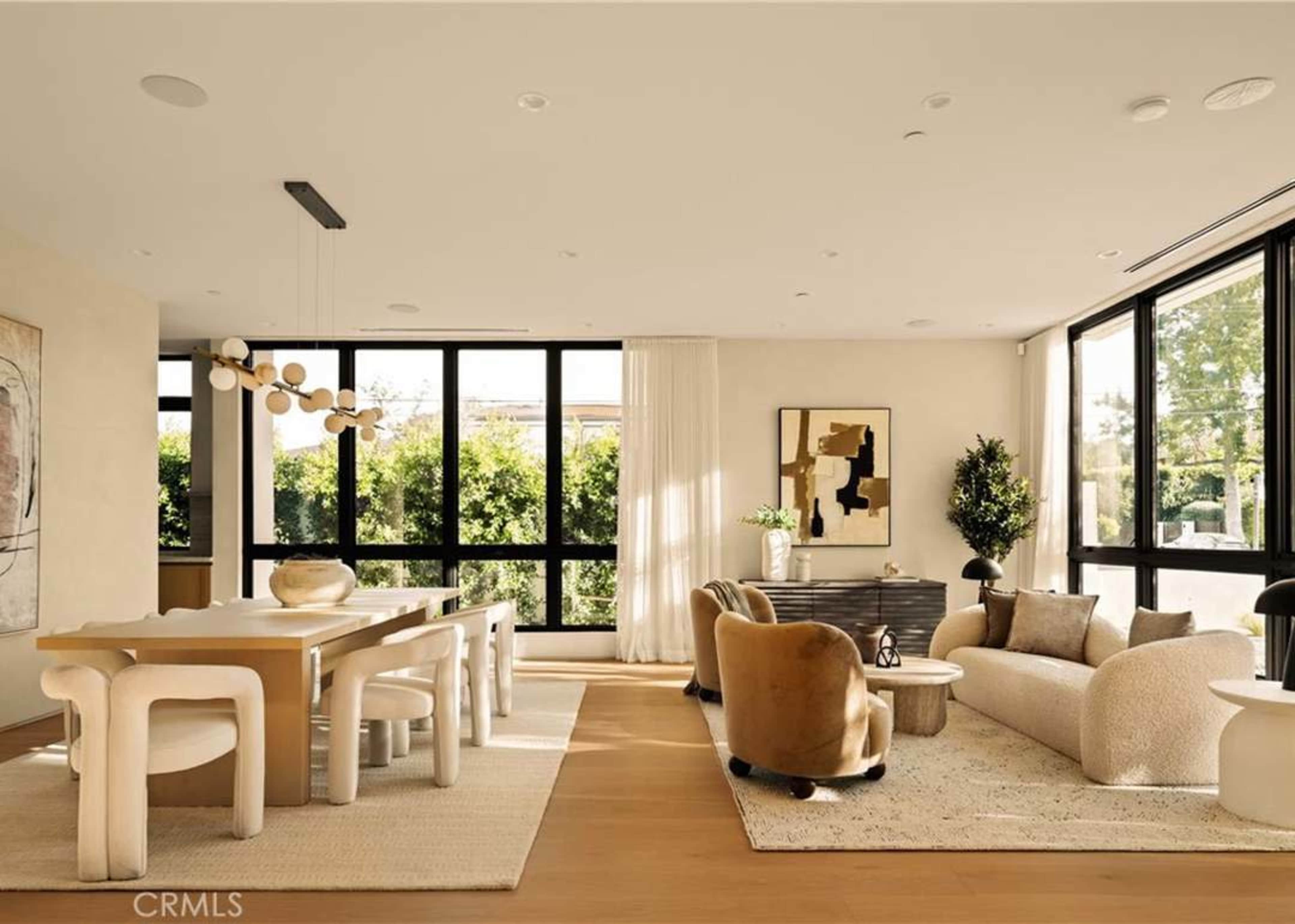 The image shows a modern living space featuring a dining area with a minimalist table, a cozy seating arrangement, large windows overlooking greenery, and neutral-toned decor.