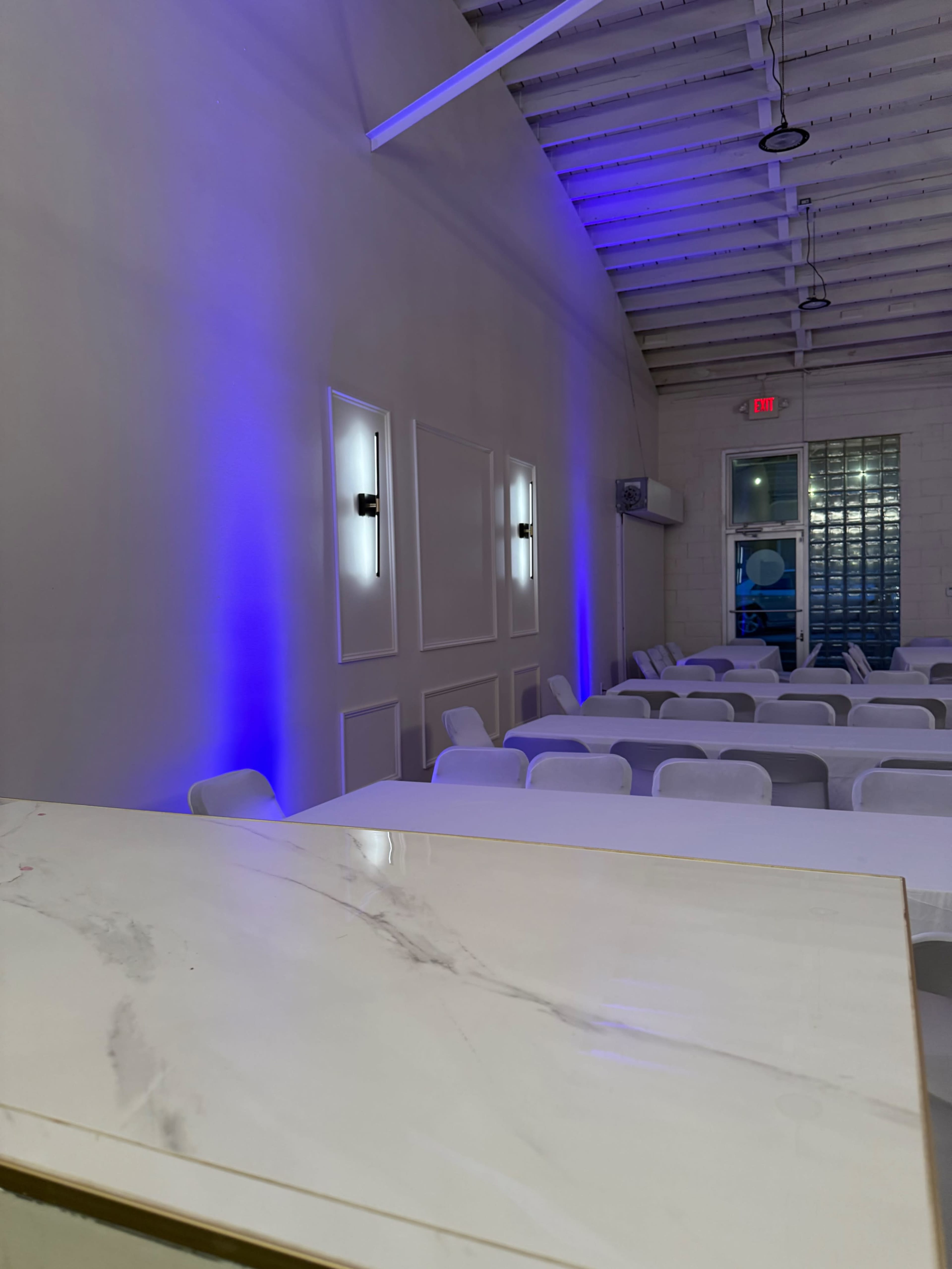 The image shows a modern event space with neatly arranged white tables and chairs, illuminated by blue lighting along the walls.