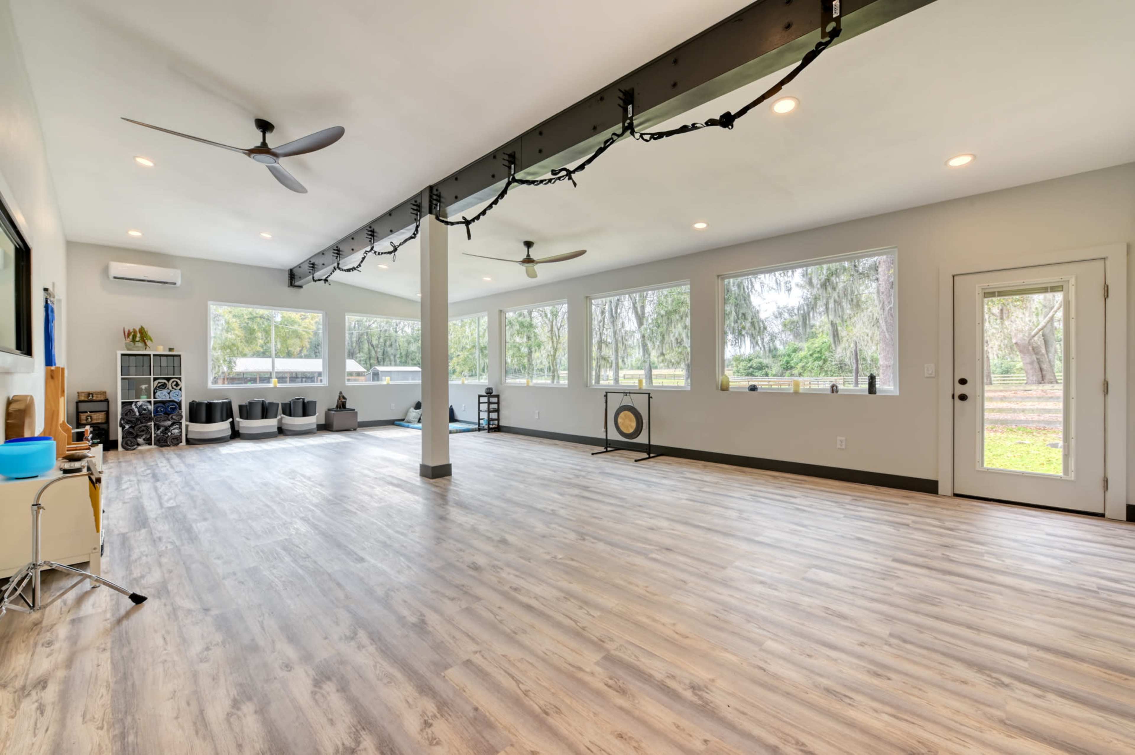 The image shows a spacious, well-lit room with large windows, light wood flooring, and a few furniture pieces arranged along the walls.