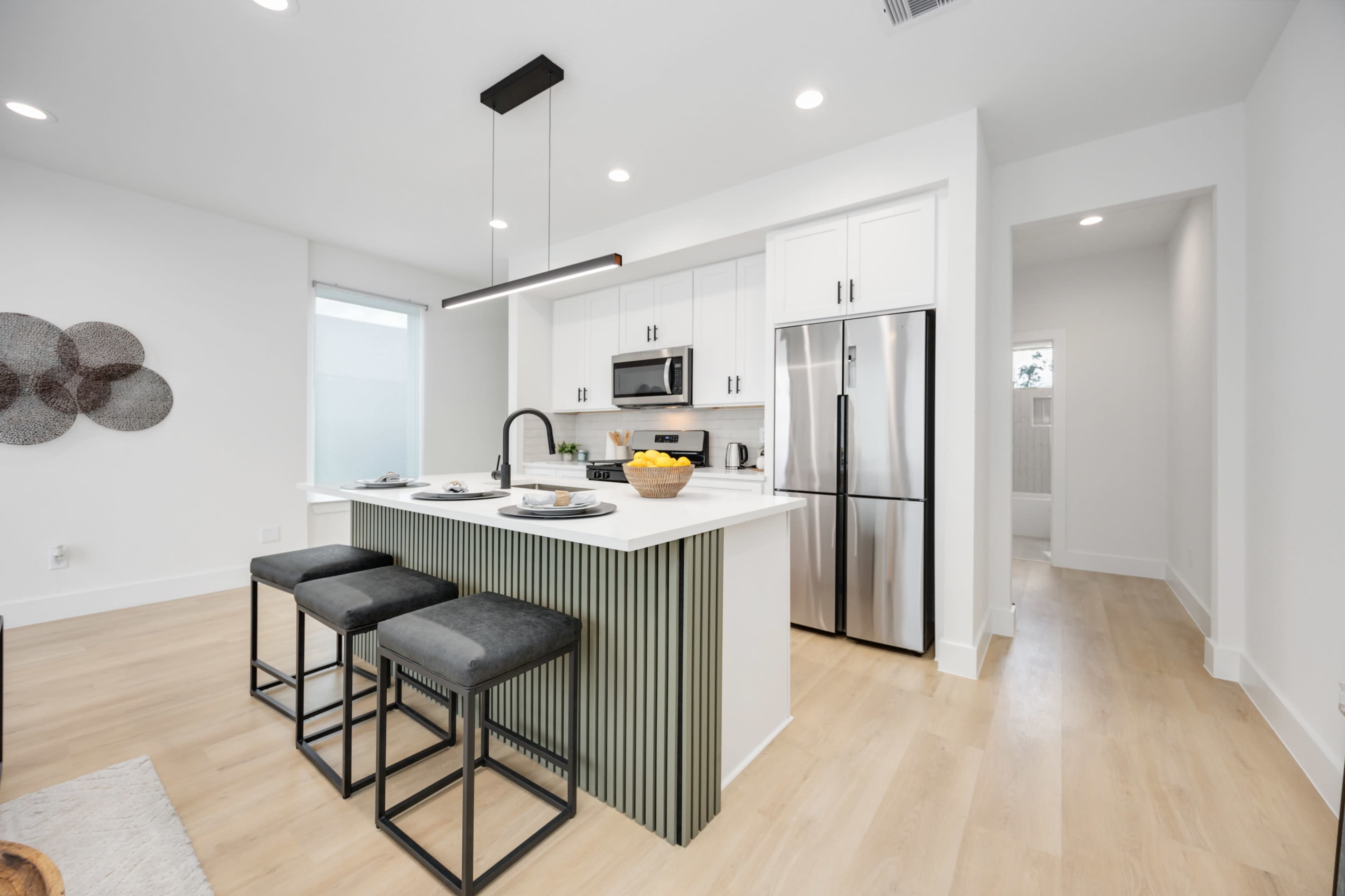 The image shows a modern kitchen with a central island featuring bar stools, stainless steel appliances, and white cabinetry.