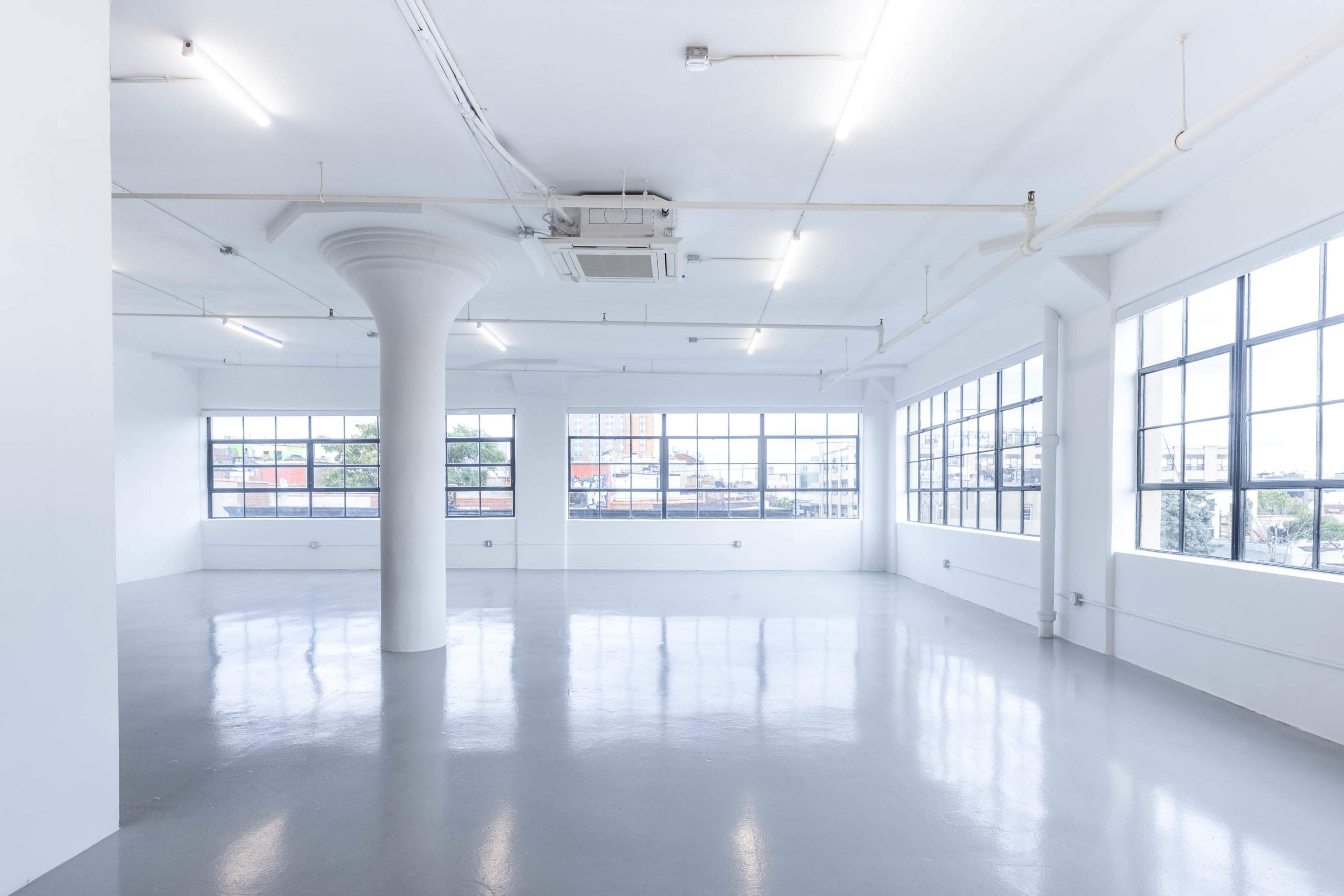 A spacious, empty room with white walls, polished gray flooring, and large windows allowing natural light to enter.