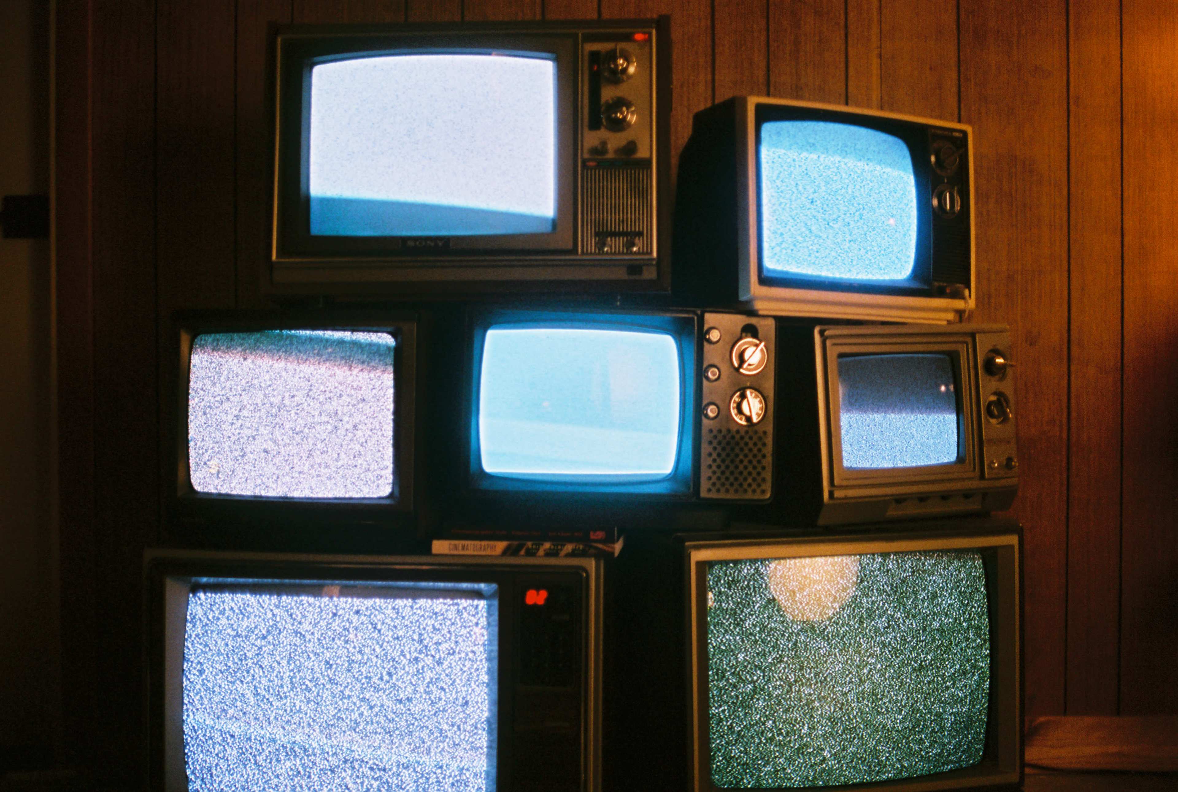 A stack of vintage televisions displaying static on their screens is placed against a wooden wall.