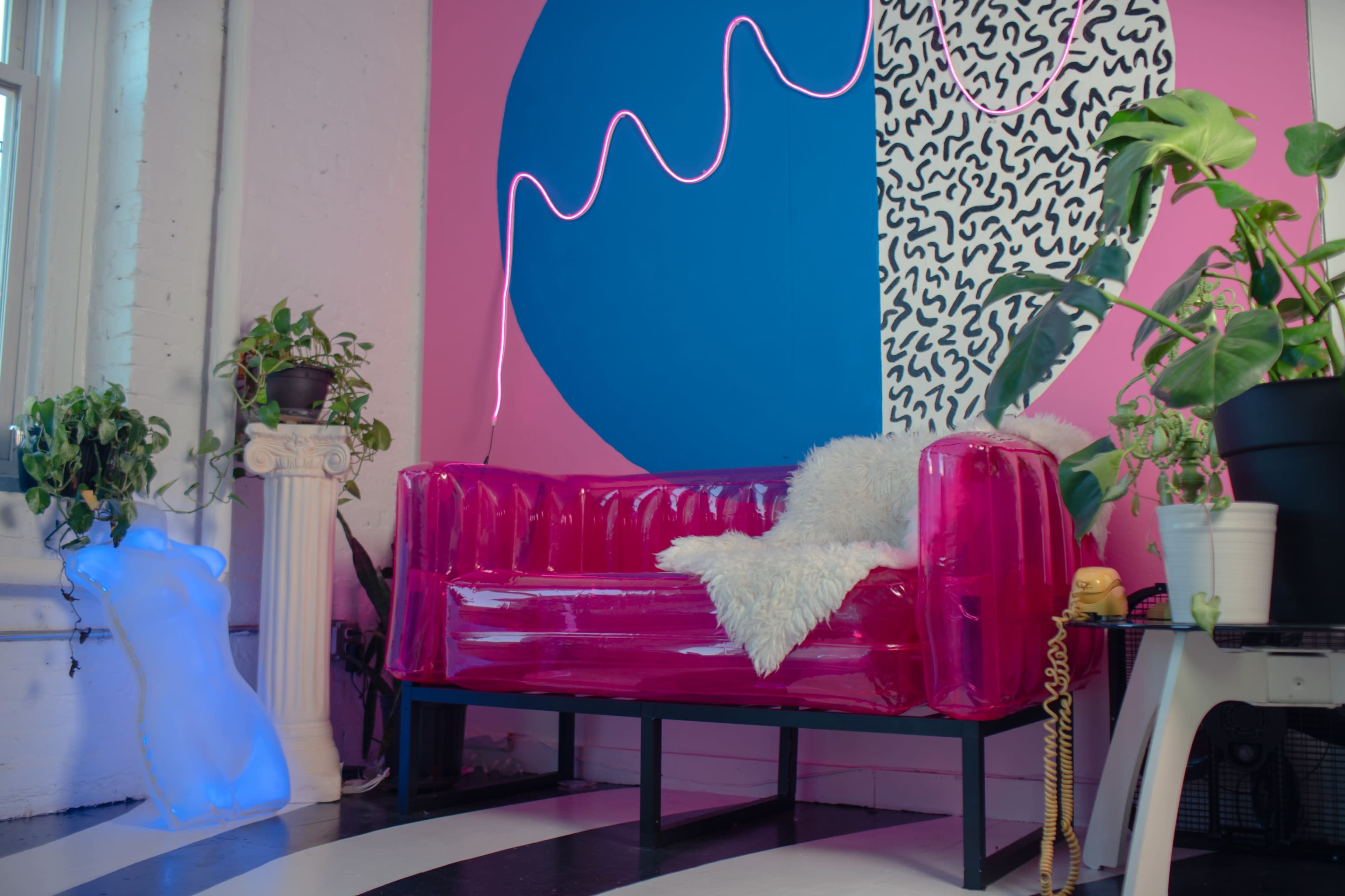 The scene features a bright pink inflatable couch with a white throw blanket, surrounded by colorful wall designs and indoor plants.