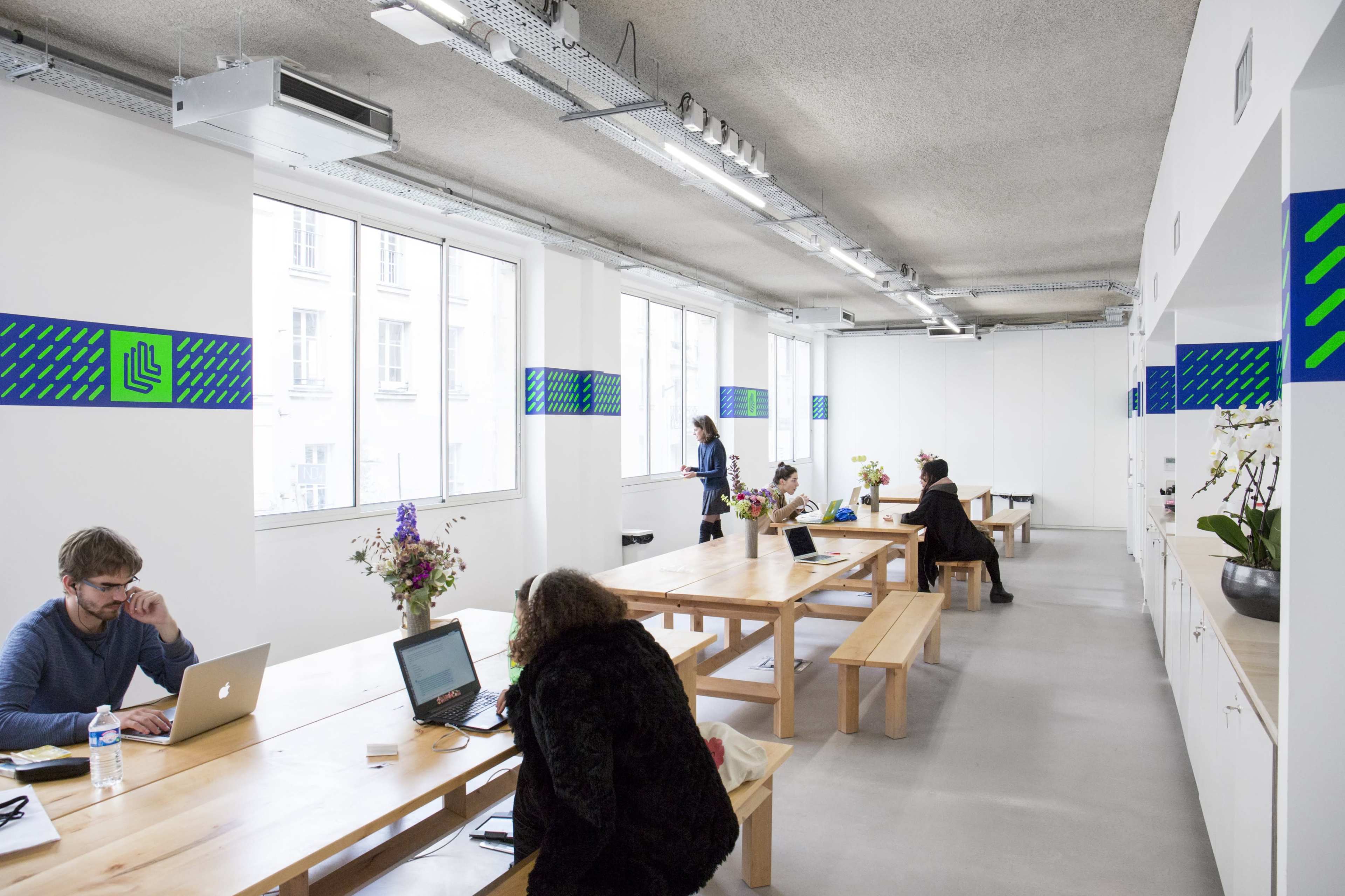 A spacious coworking space features long wooden tables, computers, and flower arrangements, with several individuals engaged in work activities.