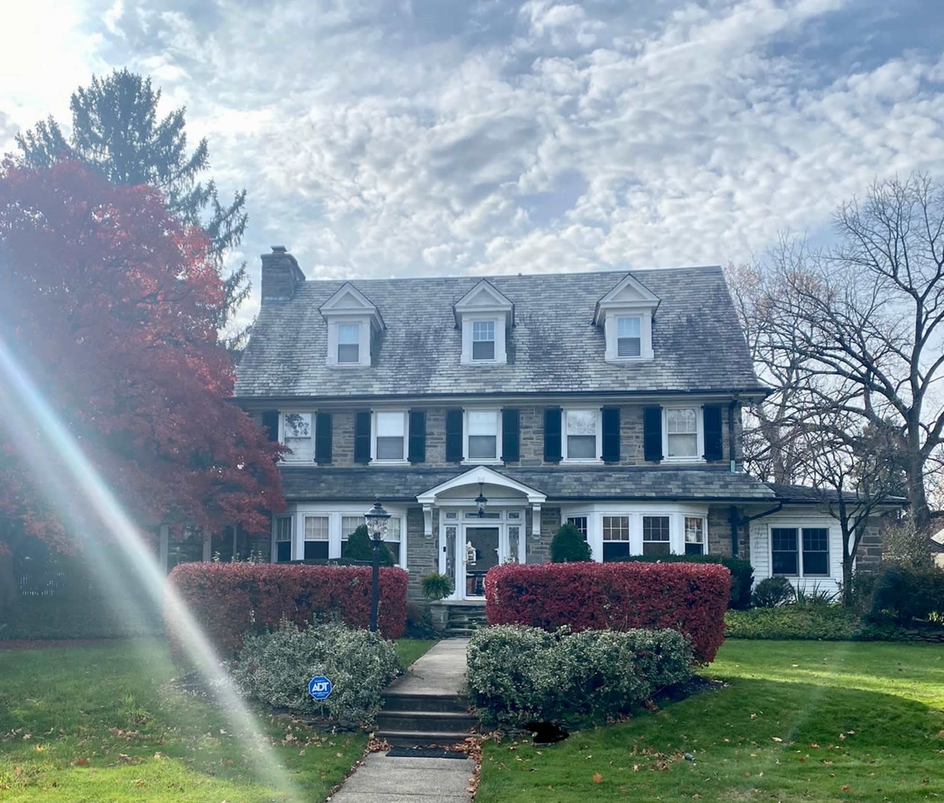 A large, gray-stone house with a gabled roof and white trim is surrounded by neatly trimmed bushes and autumn-colored trees under a cloudy sky.