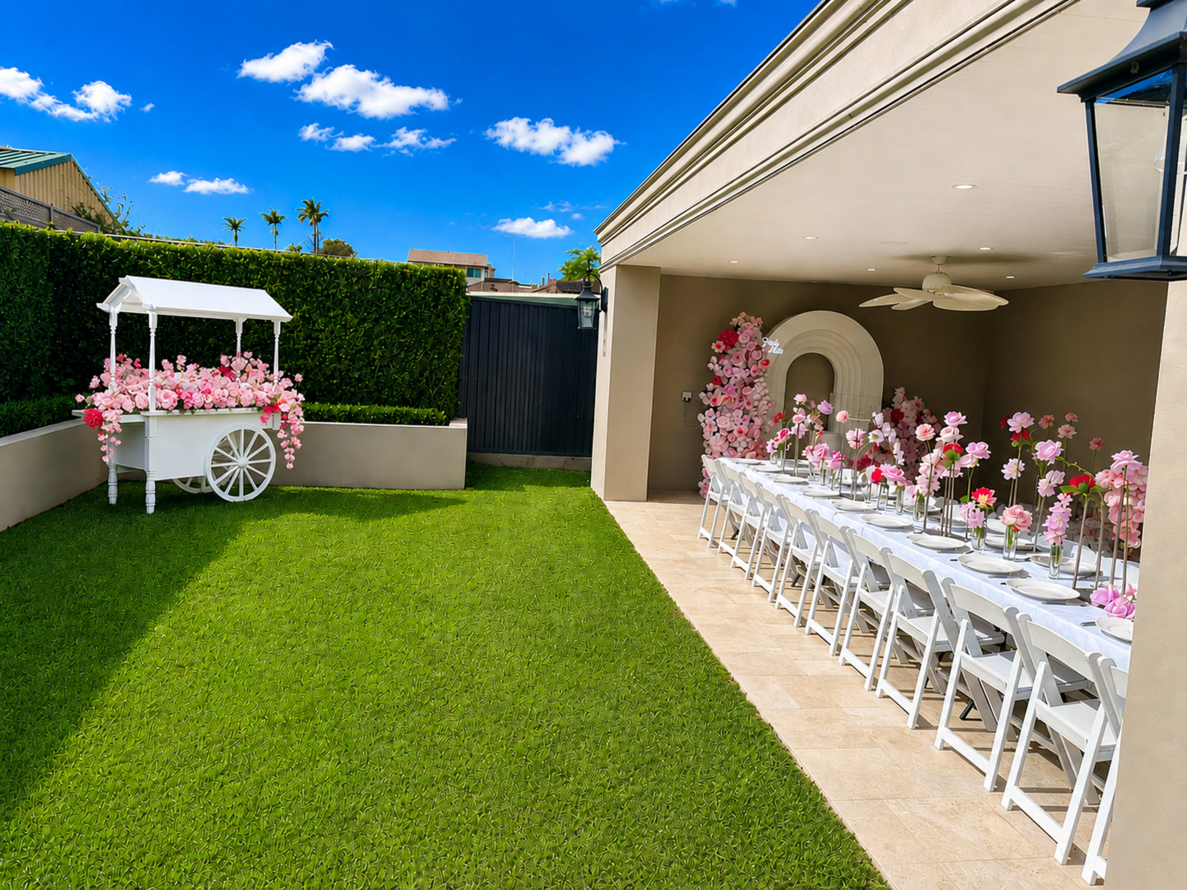 The scene features a beautifully arranged outdoor space with a long table set for an event, adorned with floral centerpieces, alongside a decorative cart filled with flowers on a well-manicured lawn.